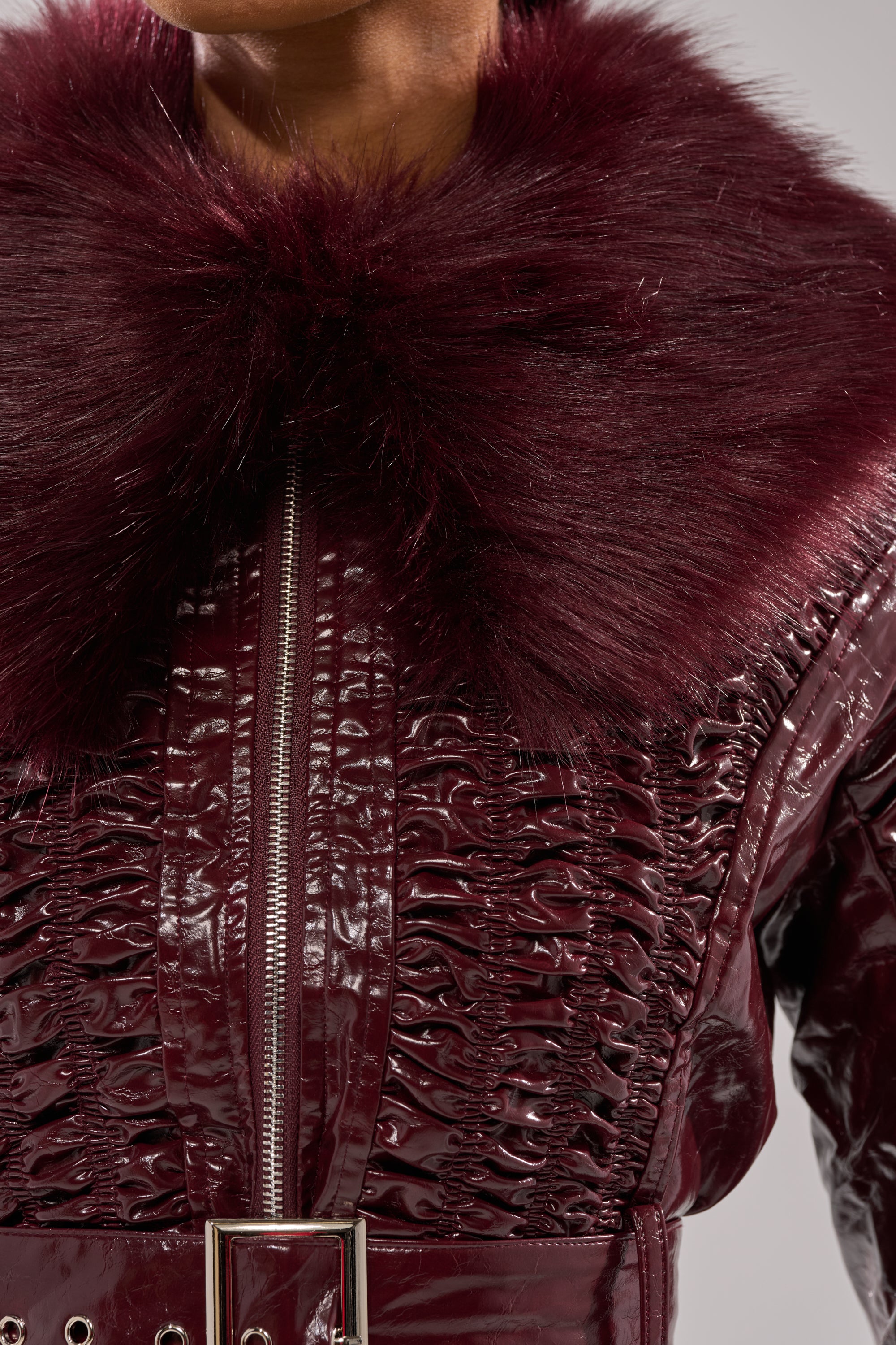 A close-up of a person wearing the IF YOU'RE DOWN FUR TRIM MOTO—a shiny, deep burgundy patent leather moto jacket with textured patterns, a large matching faux fur collar, a zipper front, and a belted waist.
