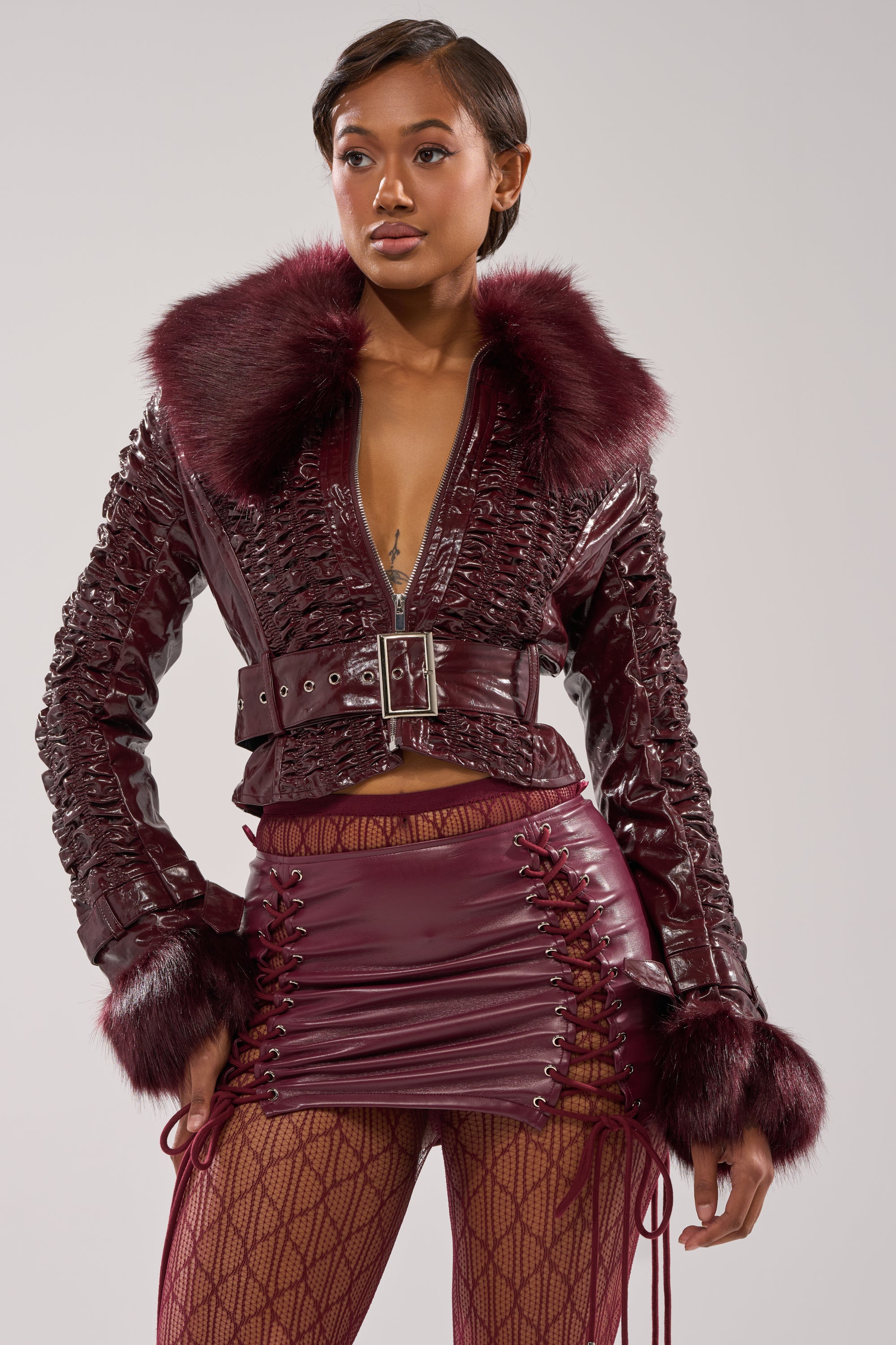 A woman models the IF YOU'RE DOWN FUR TRIM MOTO, a stylish burgundy faux fur moto jacket with belt, paired with a matching lace-up mini skirt and patterned fishnet tights. The chic winter look pops against a light gray background.