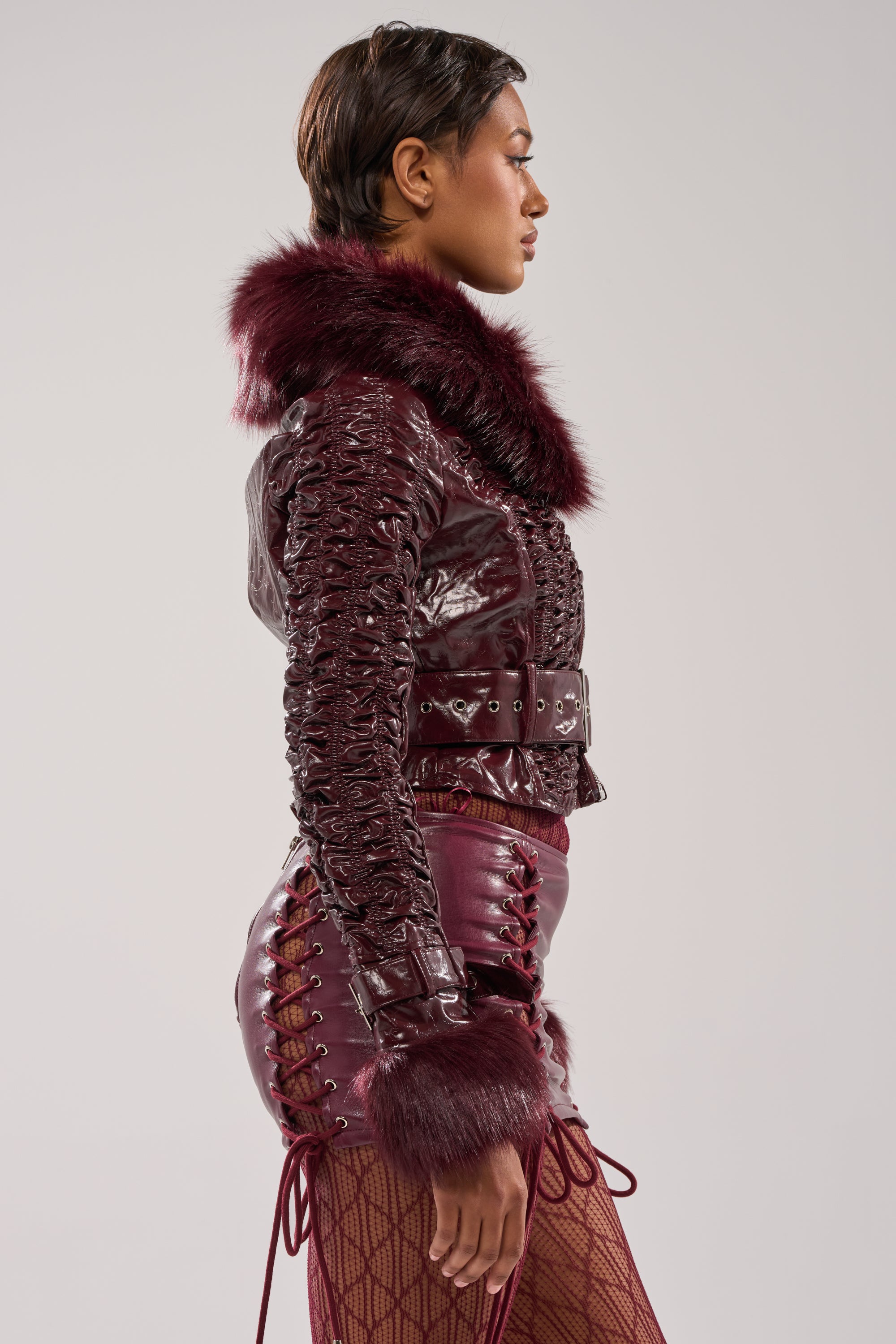 A person in profile wears the IF YOU'RE DOWN FUR TRIM MOTO jacket with lace-up details, a matching maroon mini skirt, and fishnet tights against a plain light background.