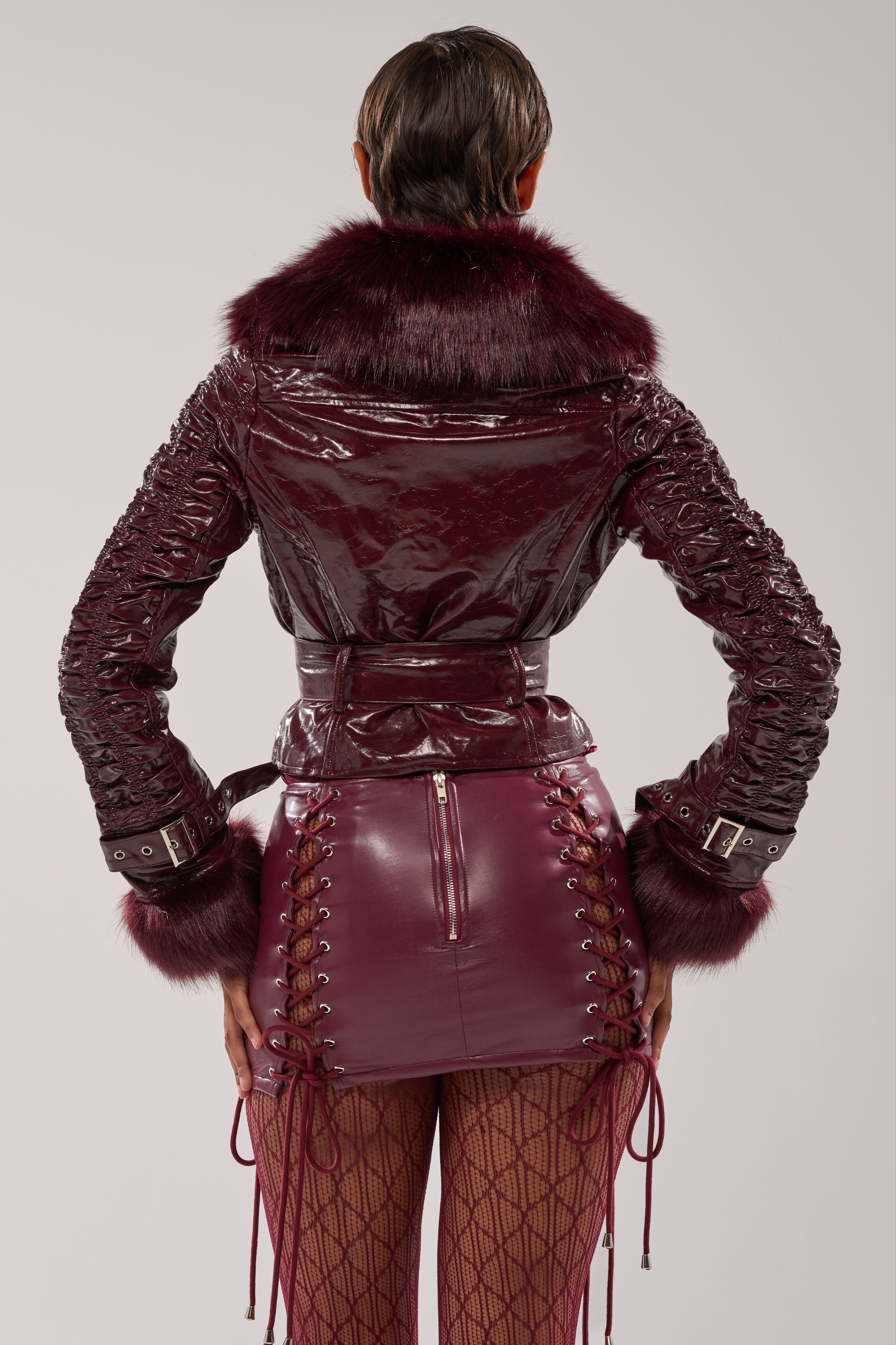 A person stands facing away, wearing the IF YOU'RE DOWN FUR TRIM MOTO— a glossy burgundy patent leather jacket with fur trim and laced sleeves—paired with a matching skirt and fishnet stockings.