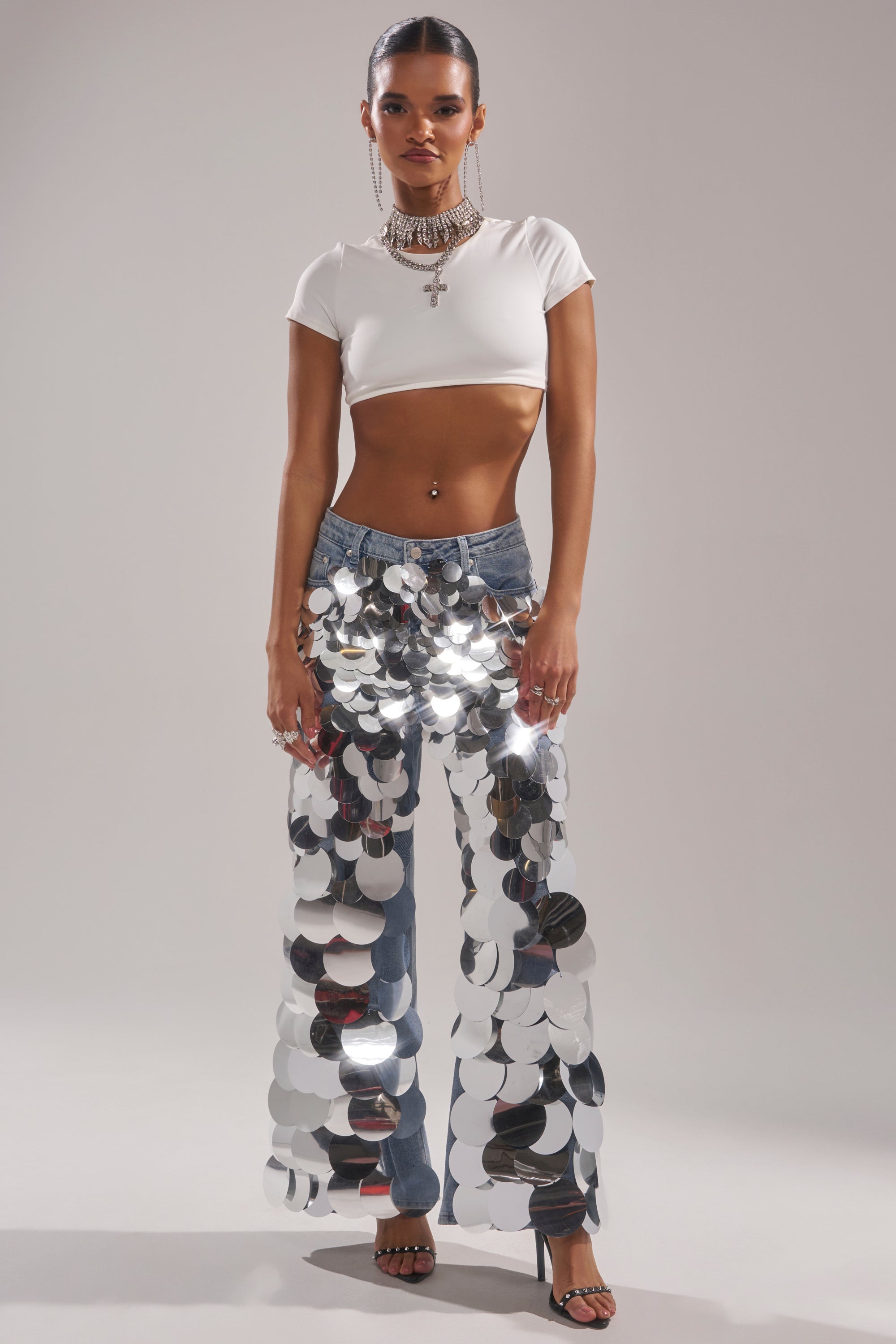 A woman wears the CHECK IT OUT SEQUIN DENIM PANT—vintage-wash blue jeans with large silver sequins—paired with a white crop top, layered necklaces, black heels, and a sleek updo, posing against a plain background.