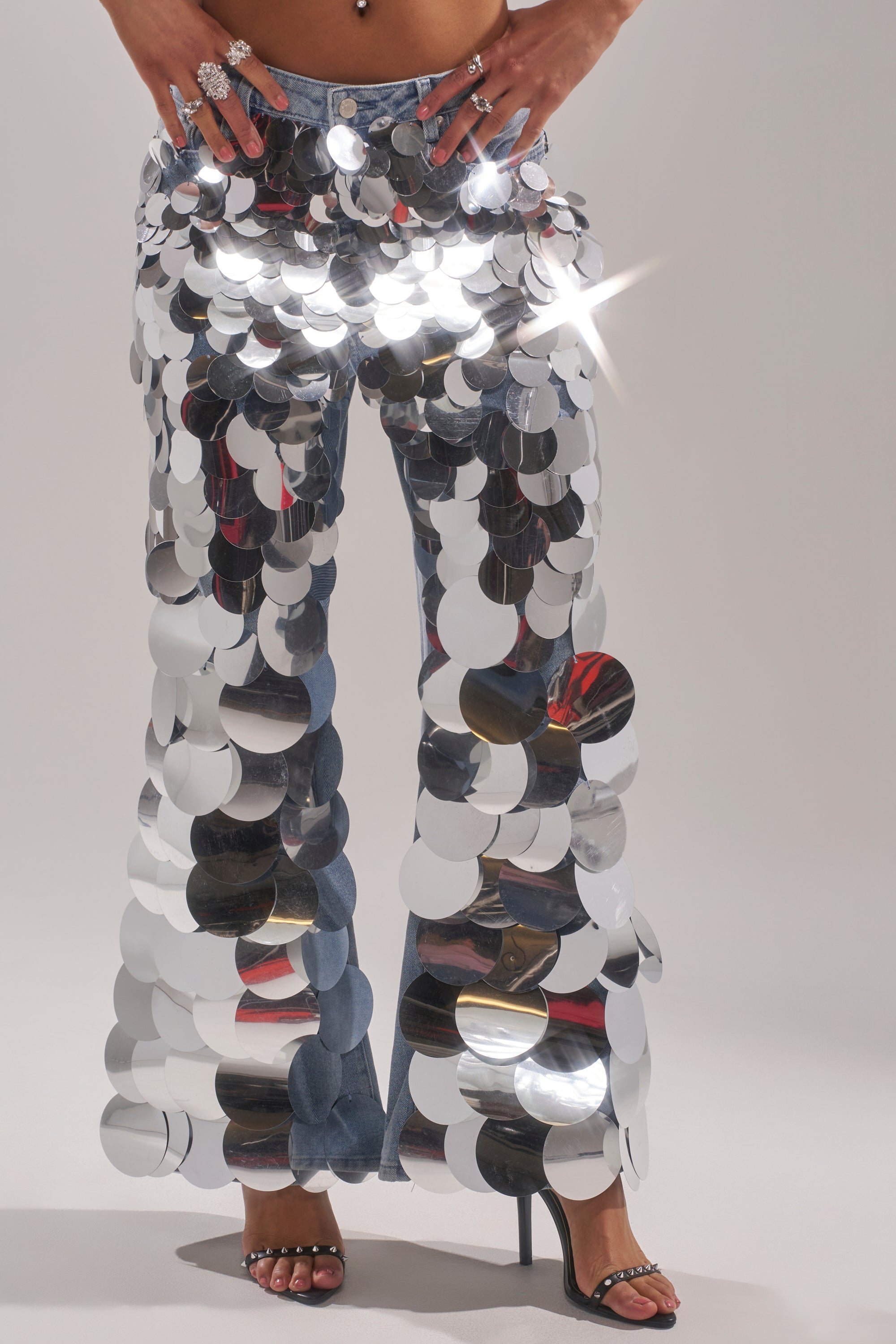 Wearing the CHECK IT OUT SEQUIN DENIM PANT in vintage-wash blue with oversized silver sequins, the person stands in high-heeled sandals. The flared legs shimmer with reflected light. Only the lower body appears.