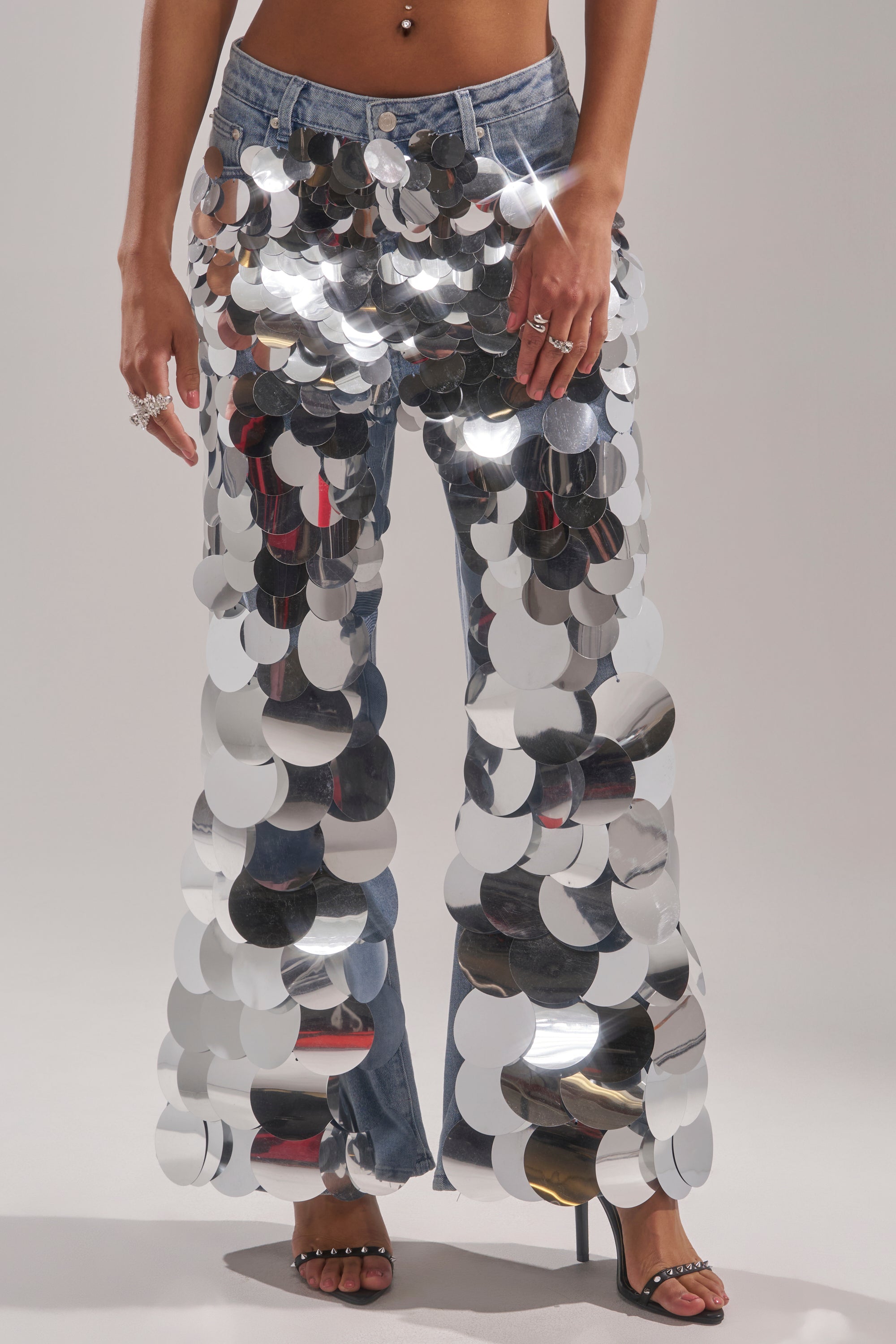A person wears the CHECK IT OUT SEQUIN DENIM PANT—vintage-wash jeans with large, shiny silver sequins—paired with heeled sandals. Only the lower body is shown, highlighting the pants' reflective, striking look.