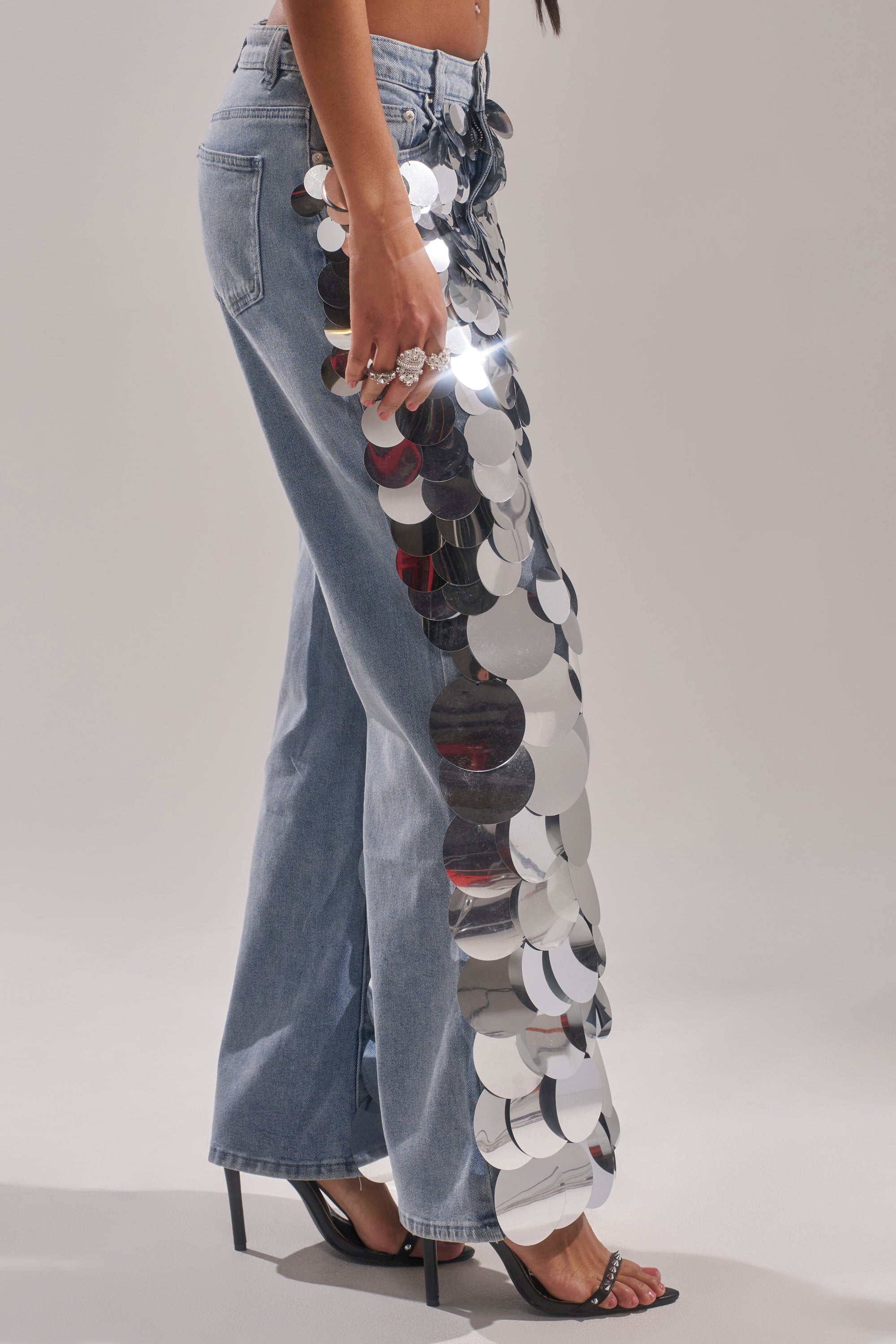A person wears the CHECK IT OUT SEQUIN DENIM PANT—vintage-wash blue jeans featuring bold silver sequins on one side—styled with black high-heeled sandals and rings, standing against a plain background.