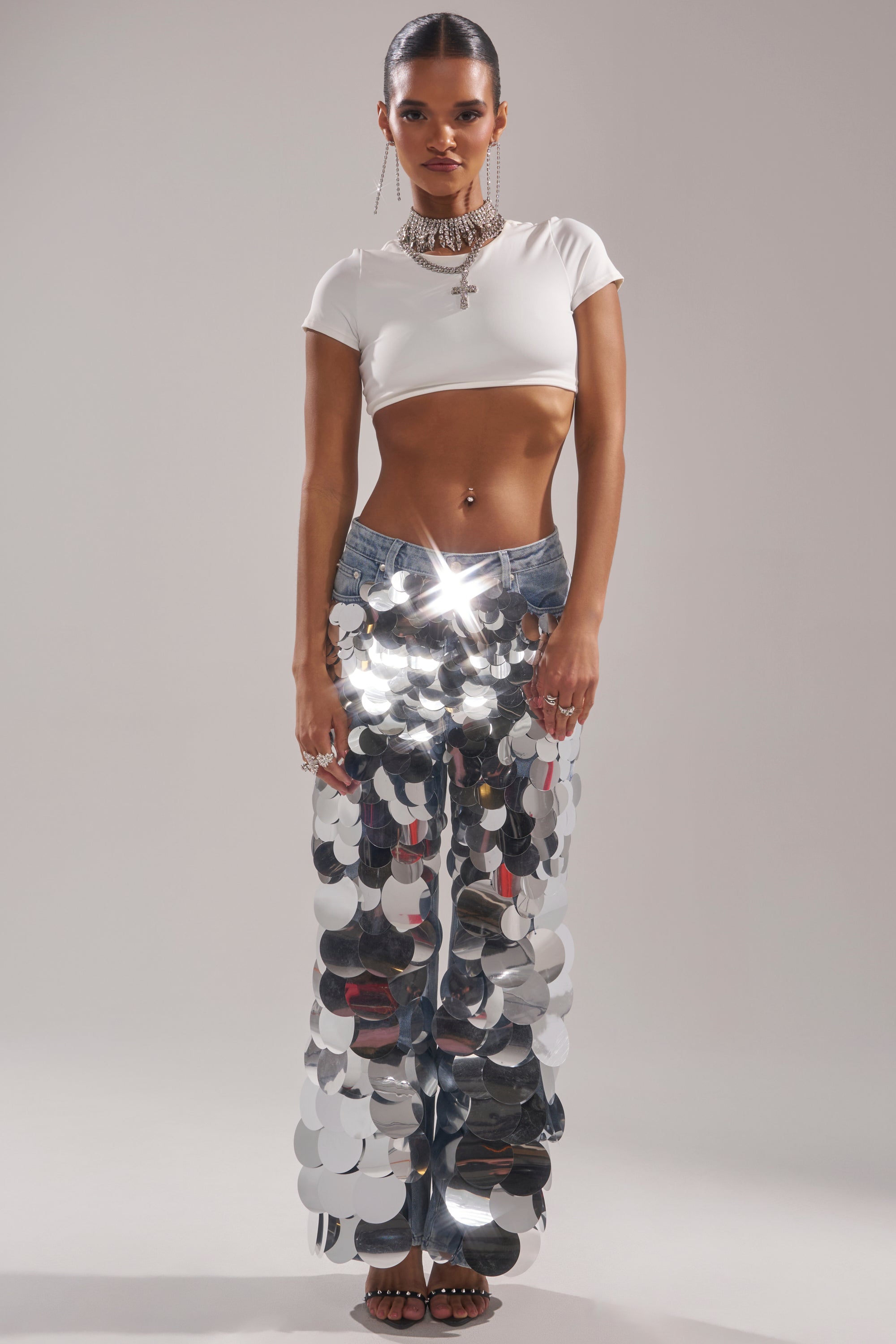 A woman poses against a plain backdrop, styling the CHECK IT OUT SEQUIN DENIM PANT with a white cropped tee, layered necklaces, and hoop earrings. The pants feature bold silver sequin discs, while her hair is slicked back for a statement look.