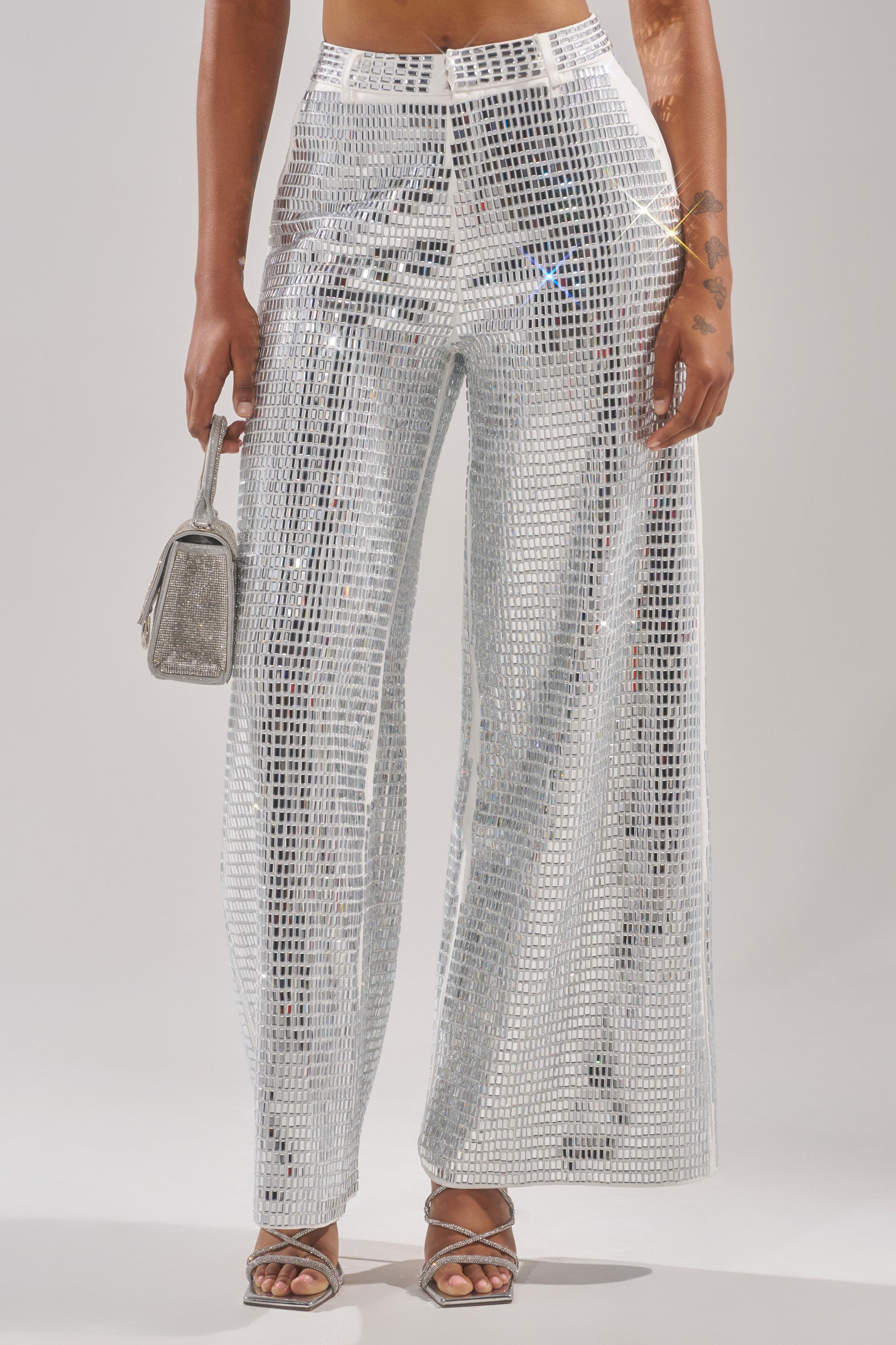 A person wears the CAN'T GET ENOUGH RHINESTONE TROUSER with strappy silver heels and holds a small glittery silver handbag. Only their lower body is visible against a plain light background. 2