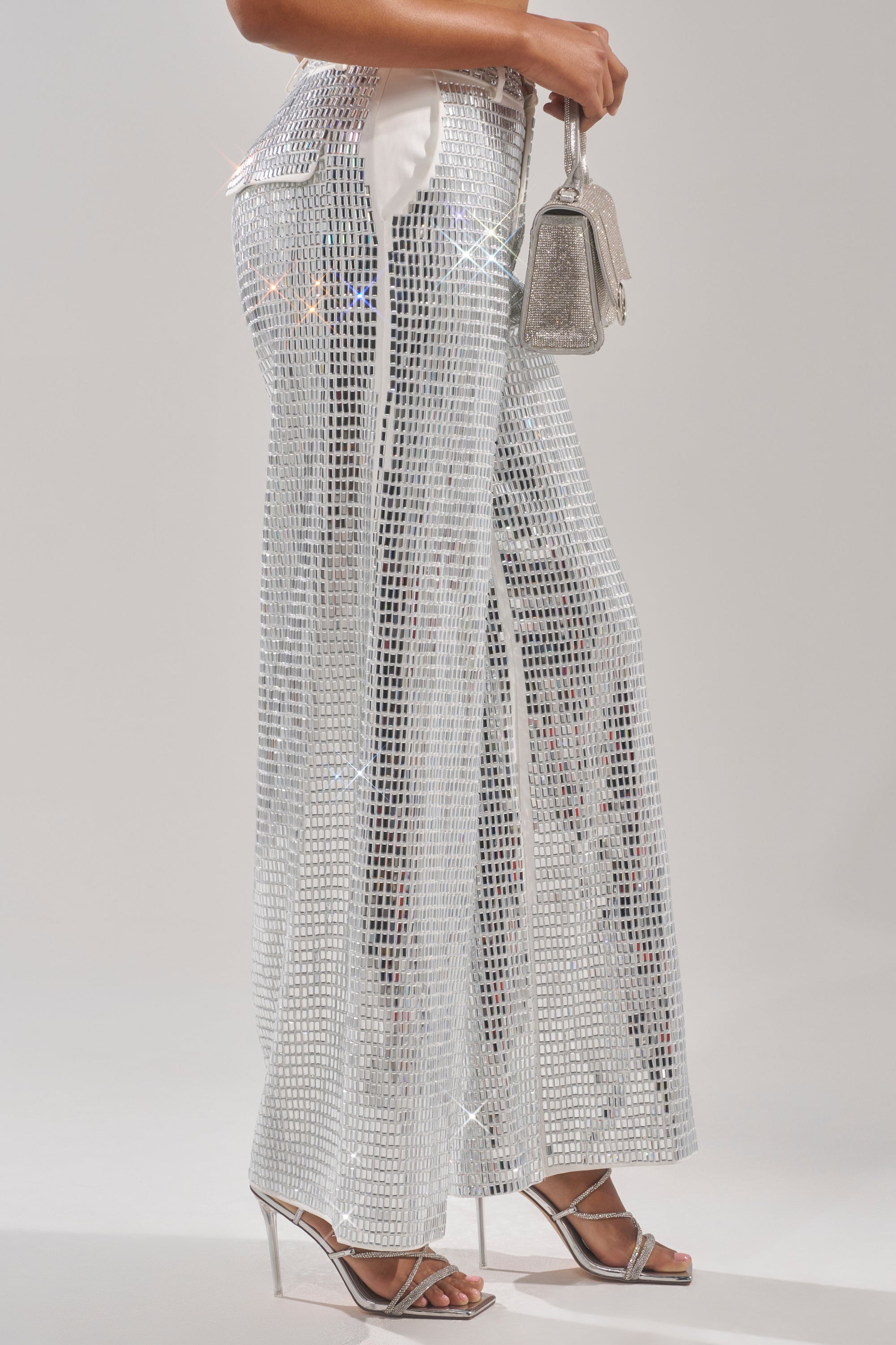 A person wears high-rise suiting with the CAN'T GET ENOUGH RHINESTONE TROUSER—wide-leg silver sequin pants—paired with silver strappy heels and a sparkly silver handbag against a plain, light-colored background. 4