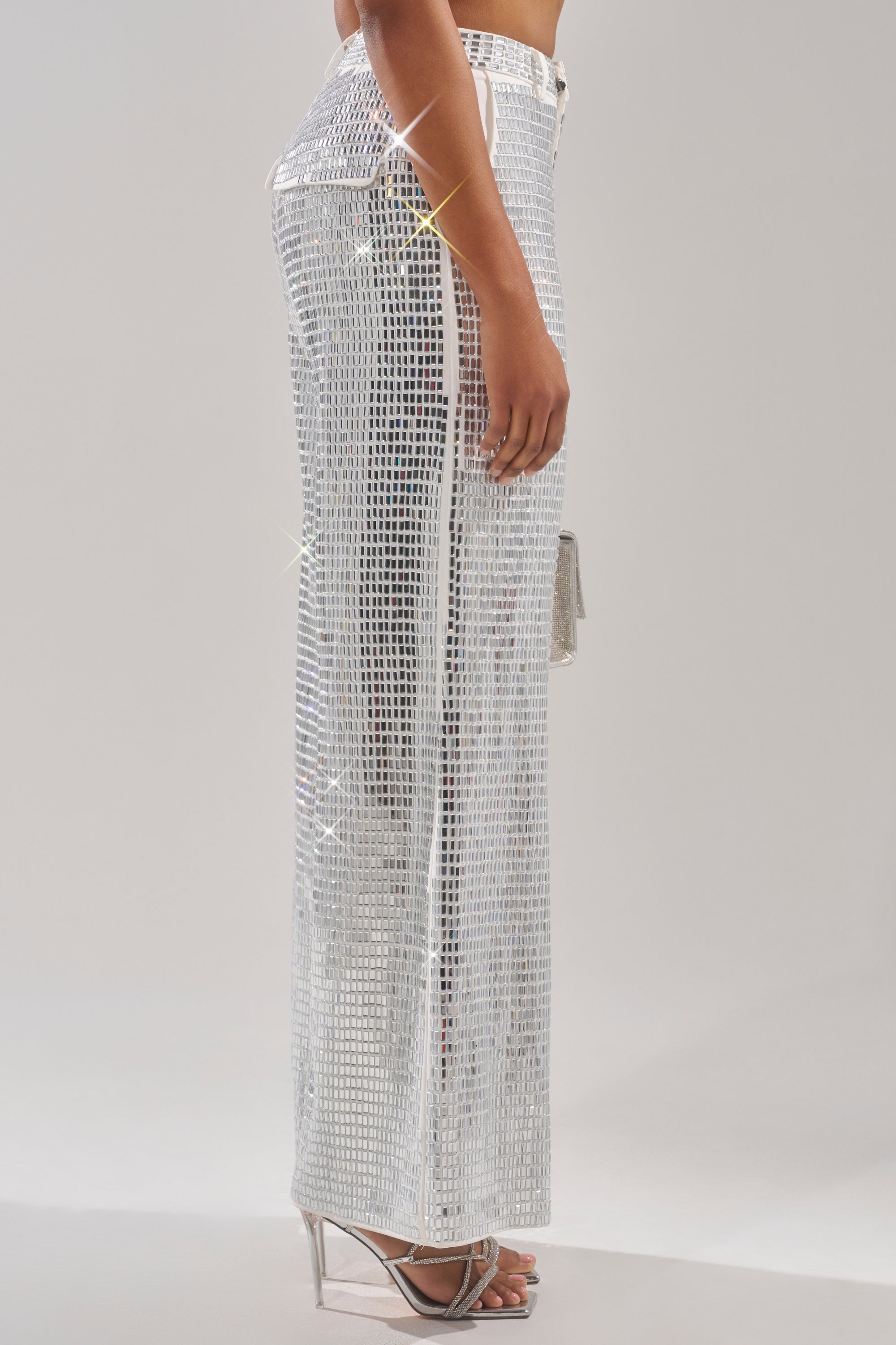 A person stands in profile wearing the CAN'T GET ENOUGH RHINESTONE TROUSER—wide-leg, silver sequin pants that reflect light—paired with silver high-heeled sandals and a small silver purse, set against a plain light background. 5