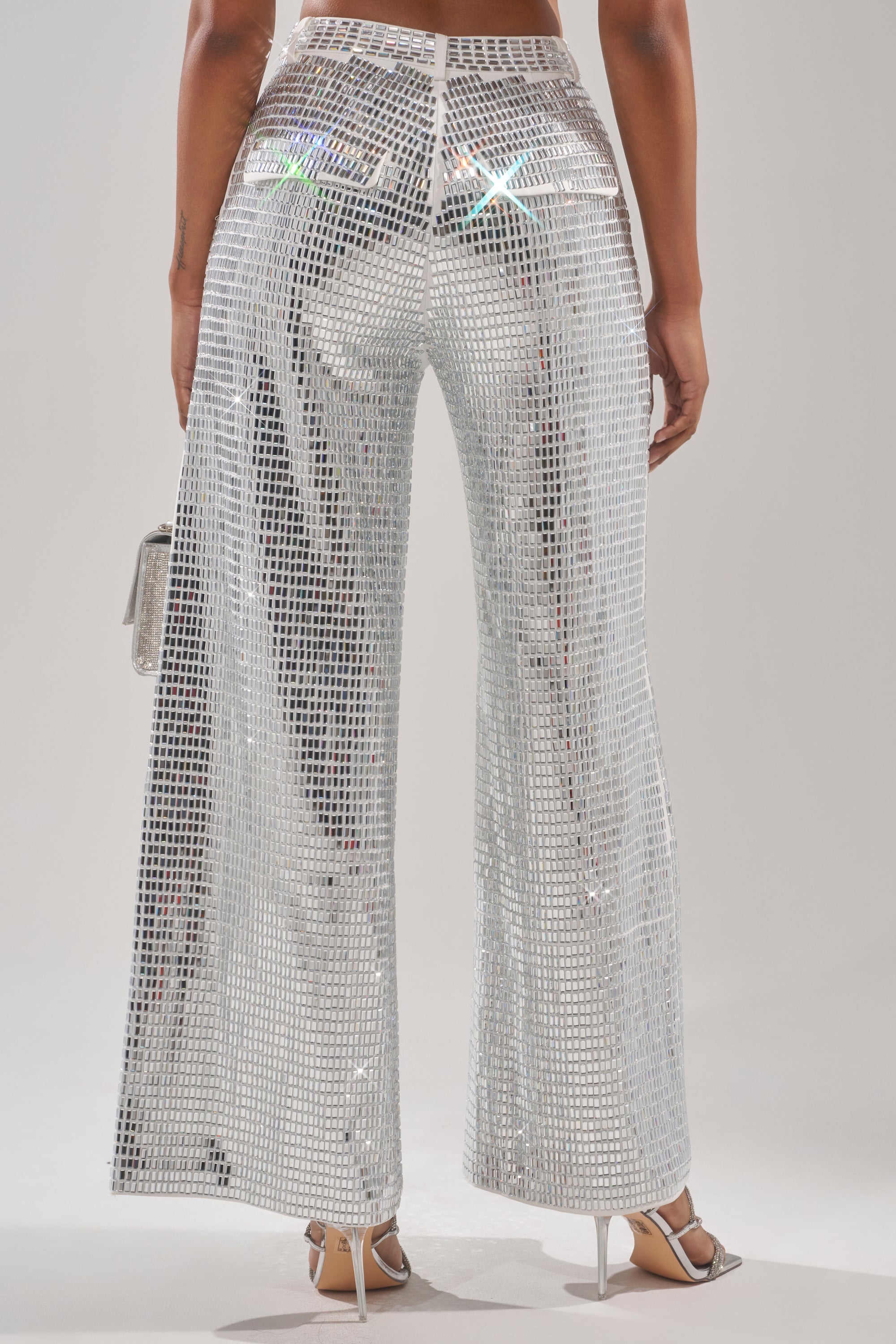 A person is seen from behind wearing the CAN'T GET ENOUGH RHINESTONE TROUSER—high-rise, wide-leg pants adorned with reflective silver square sequins—styled with white high-heeled sandals and a small silver bag. 6