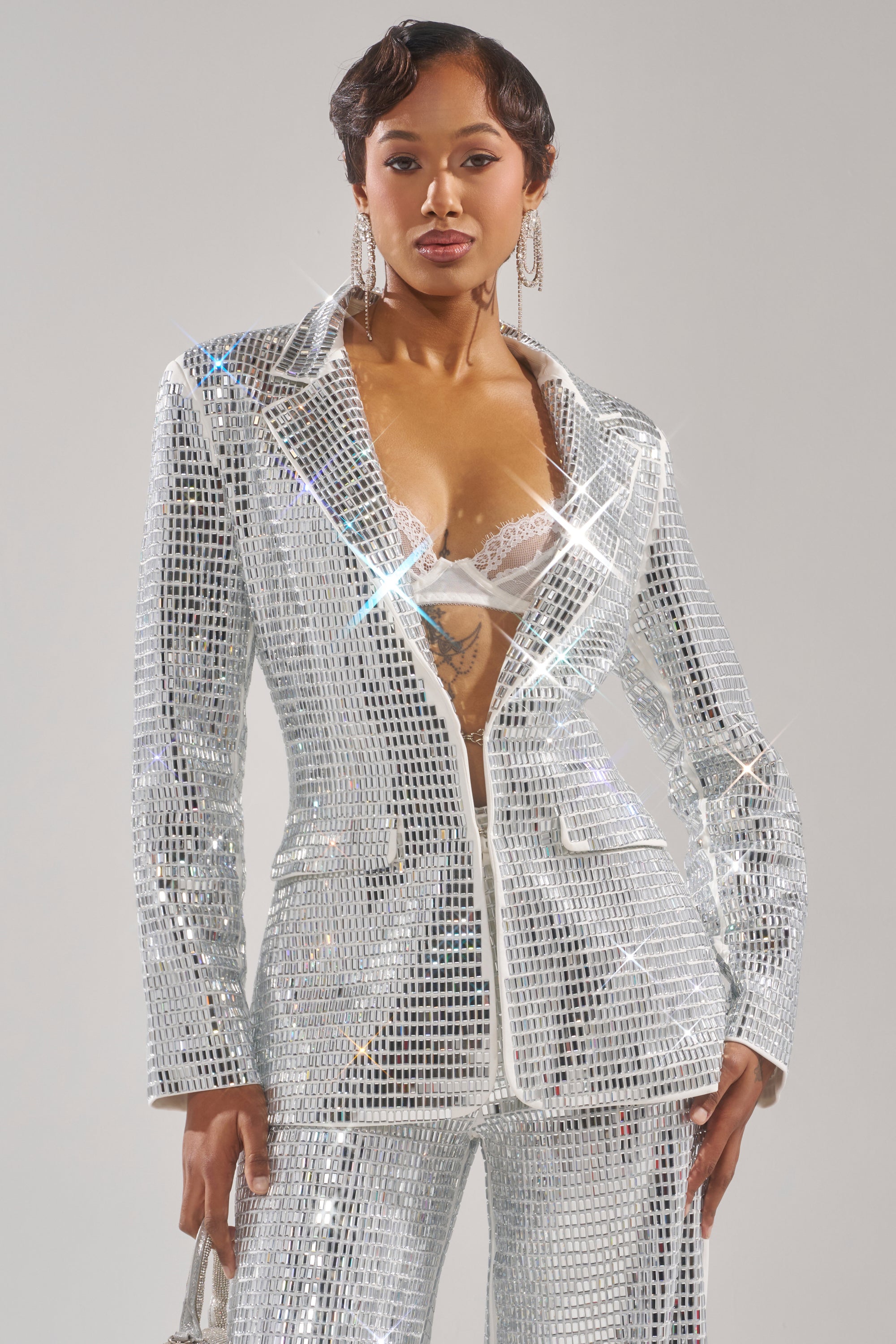 A woman poses confidently in the CAN'T GET ENOUGH RHINESTONE BLAZER, featuring a sparkling silver finish, tailored fit, and deep neckline that reveals a white lace bra. She accessorizes with statement earrings and sports short, styled hair. 0