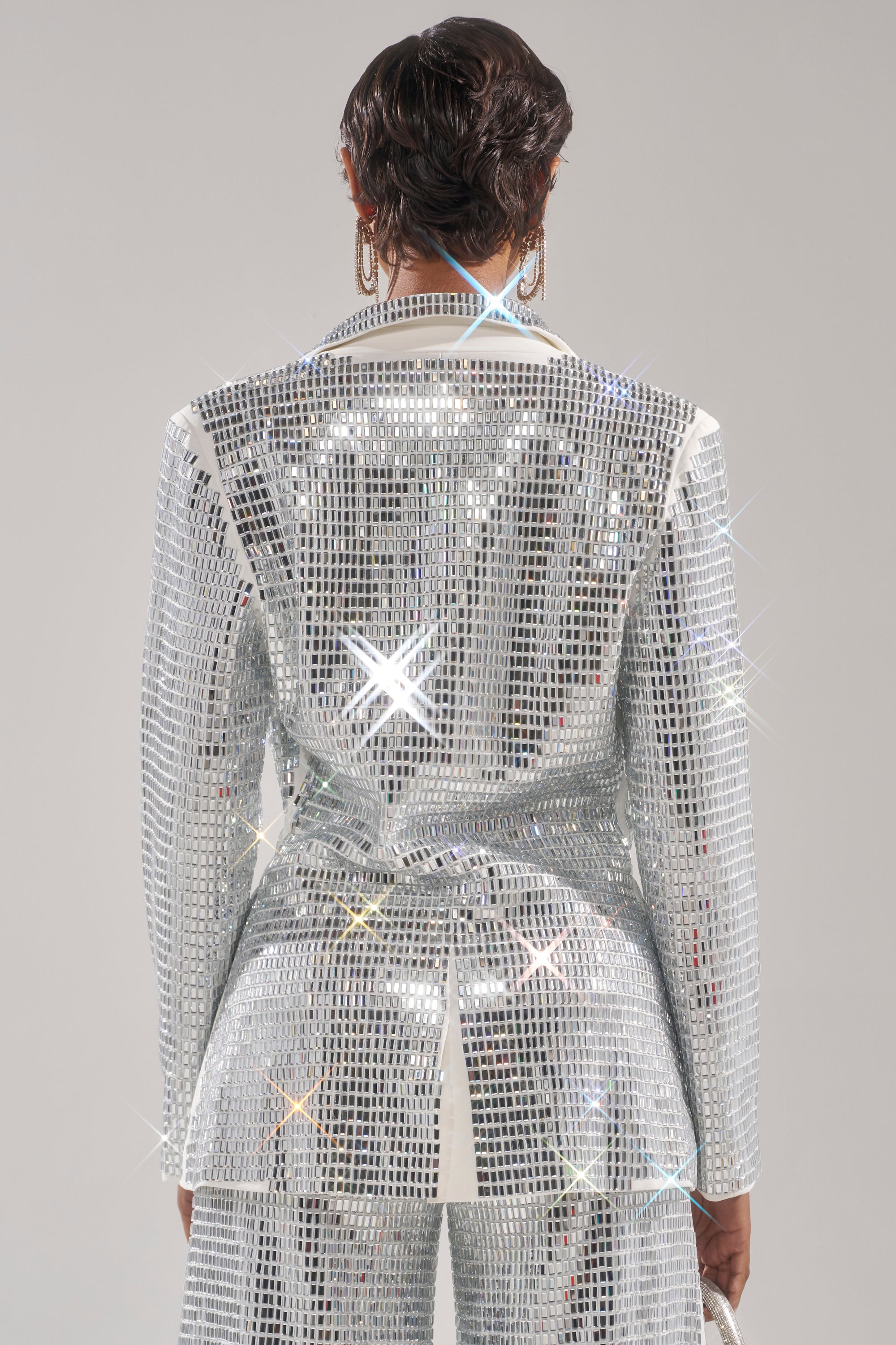 A person stands facing away, wearing the CAN'T GET ENOUGH RHINESTONE BLAZER—an oversized silver piece that shines with star-like sparkles against a plain, neutral background, highlighting its dazzling mirror effect. 4