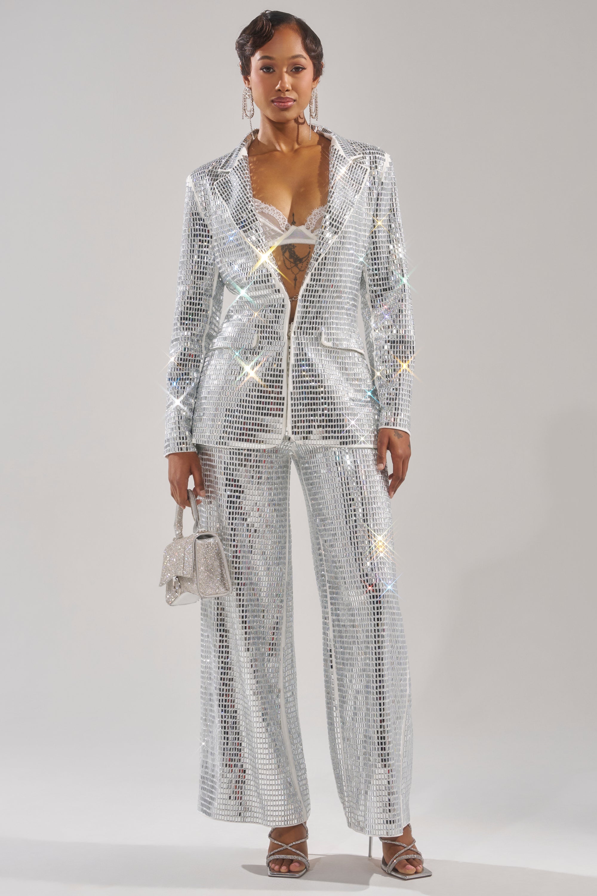 A woman stands confidently in the CAN'T GET ENOUGH RHINESTONE BLAZER, featuring sparkling silver rhinestones and a deep neckline, paired with a small silver handbag and high-heeled sandals against a plain light background. 5