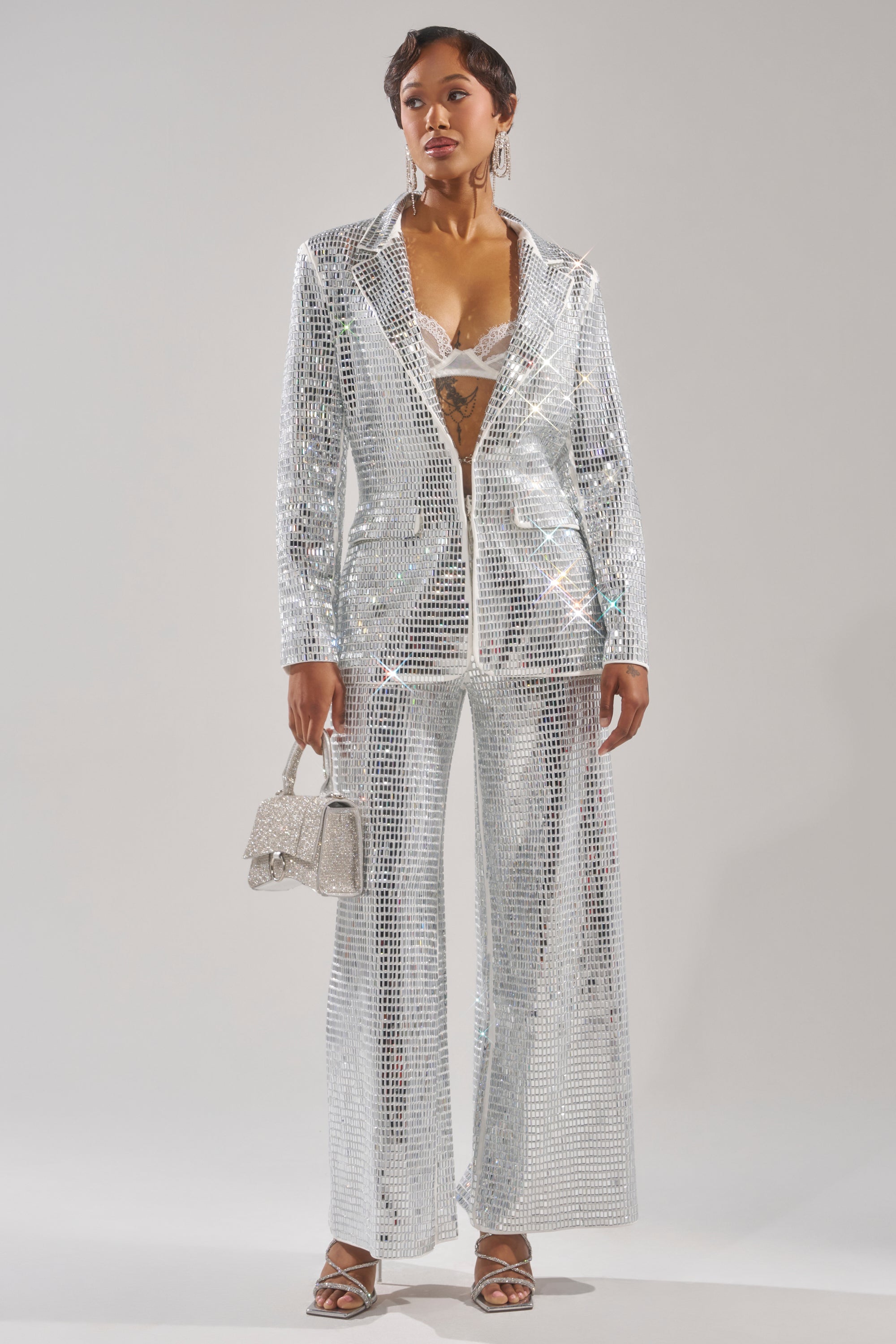 A person models the CAN'T GET ENOUGH RHINESTONE BLAZER over a white bralette and silver sequin pants, accessorizing with a sparkling handbag and silver heels against a plain, light background for a glamorous effect. 7