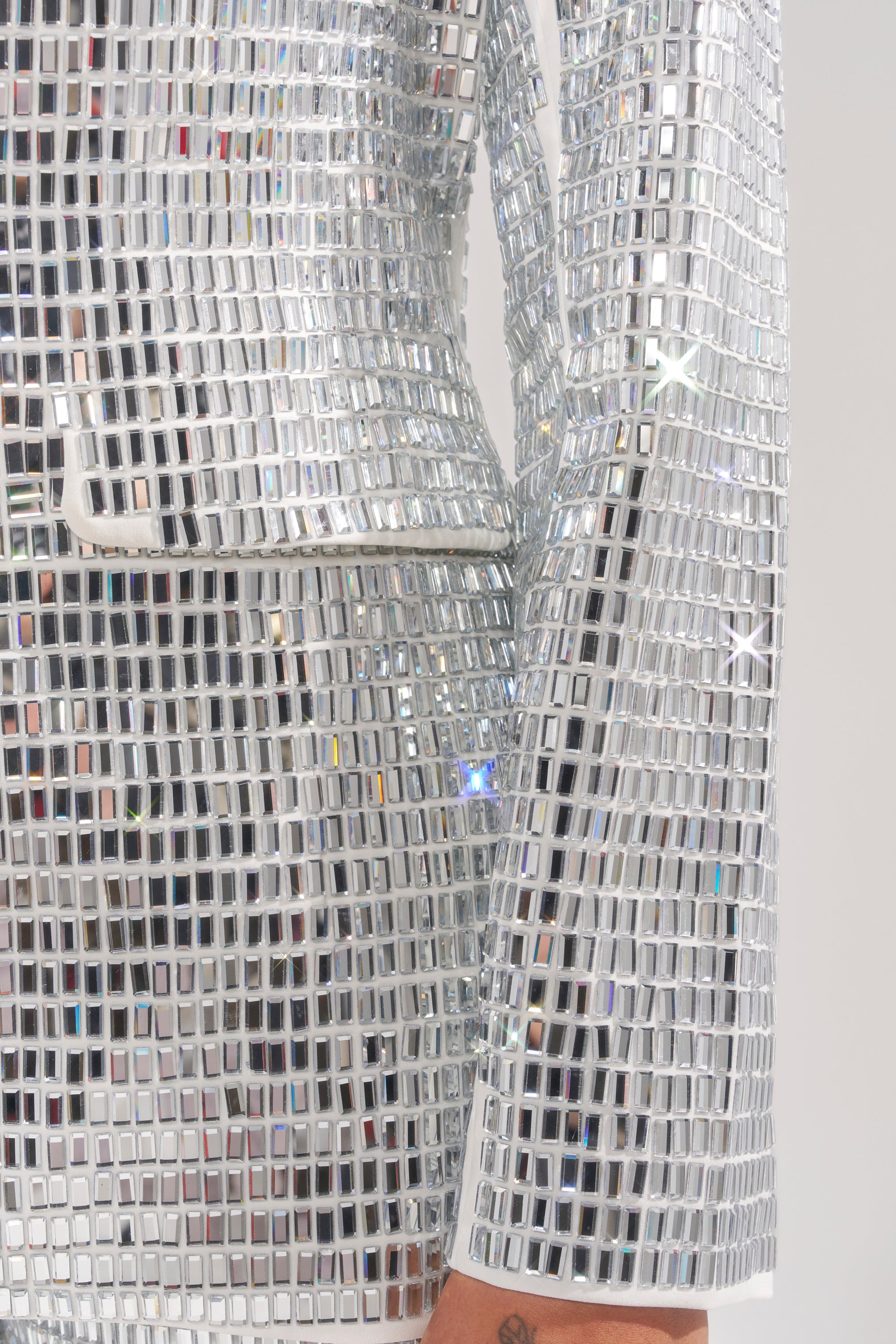 A close-up of an arm in the CAN'T GET ENOUGH RHINESTONE BLAZER, adorned with reflective silver mirror tiles. The hand and pocket are partially visible against a plain, light background. 8