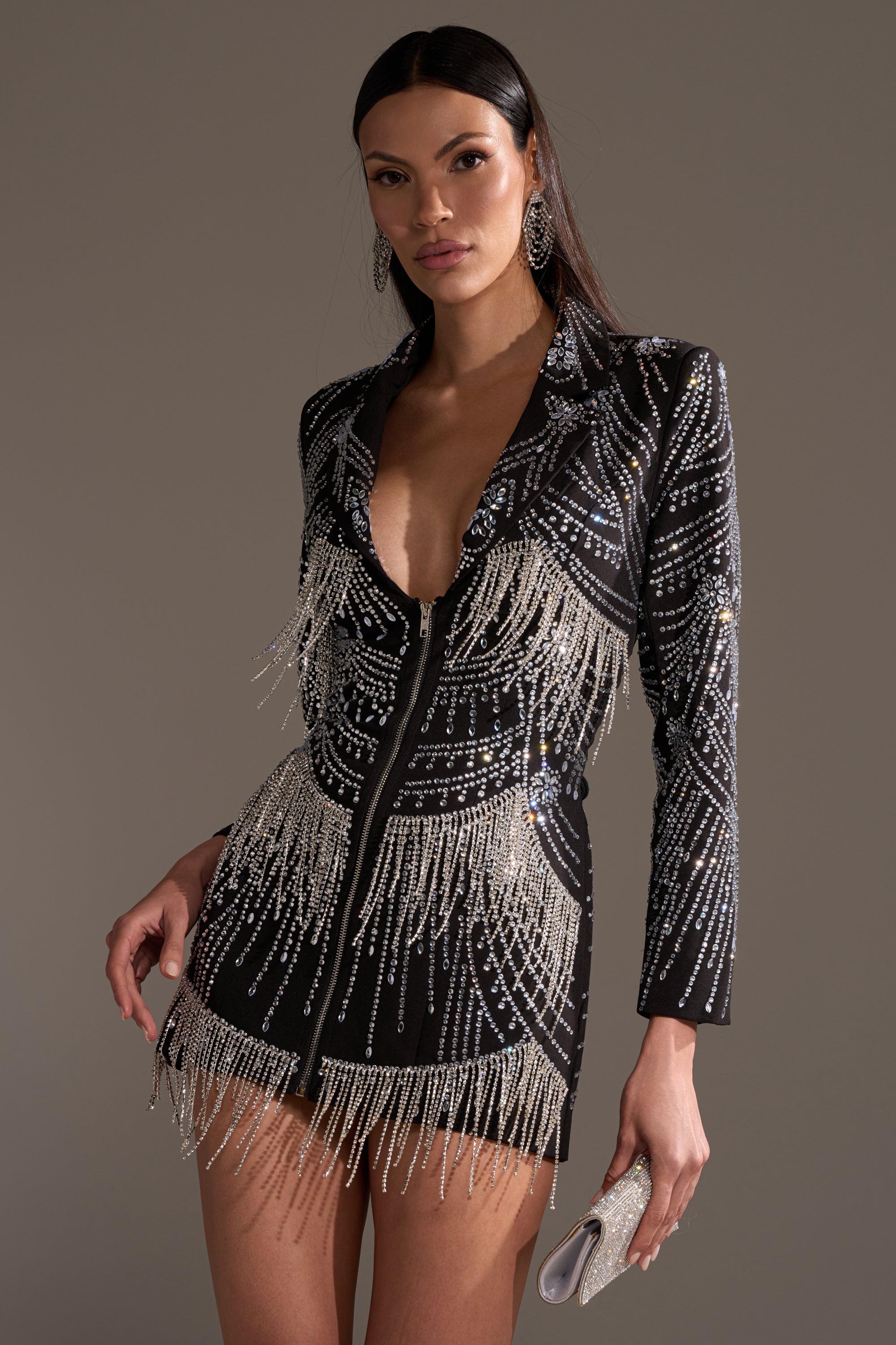 A woman in the NOT YOUR BABE RHINESTONE BLAZER DRESS with sparkling silver fringe and corset-style lace-up accents holds a small silver clutch against a plain background.