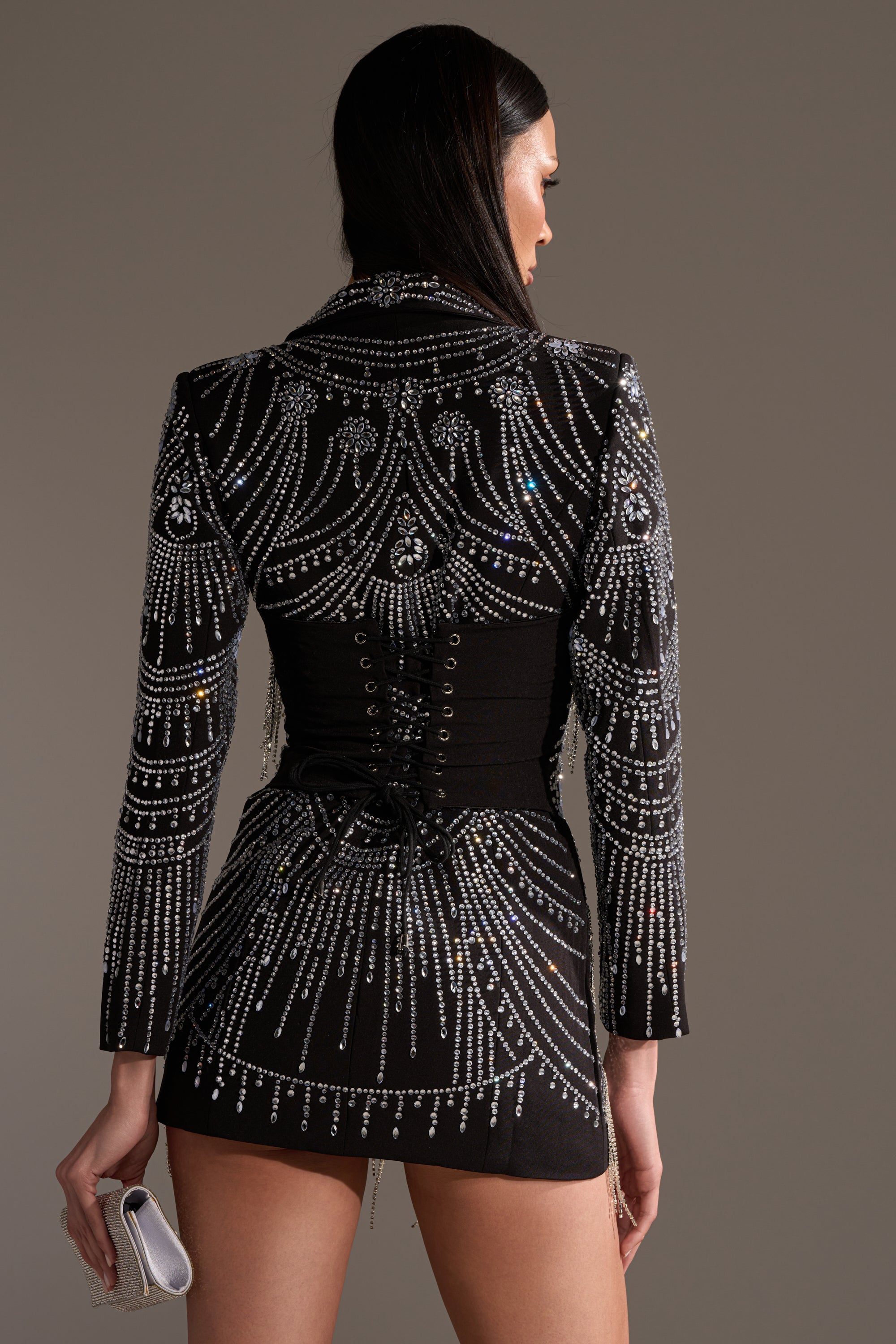 A woman faces away, wearing the NOT YOUR BABE RHINESTONE BLAZER DRESS with silver beadwork. She holds a silver clutch in her left hand, and her dark straight hair cascades down her back.