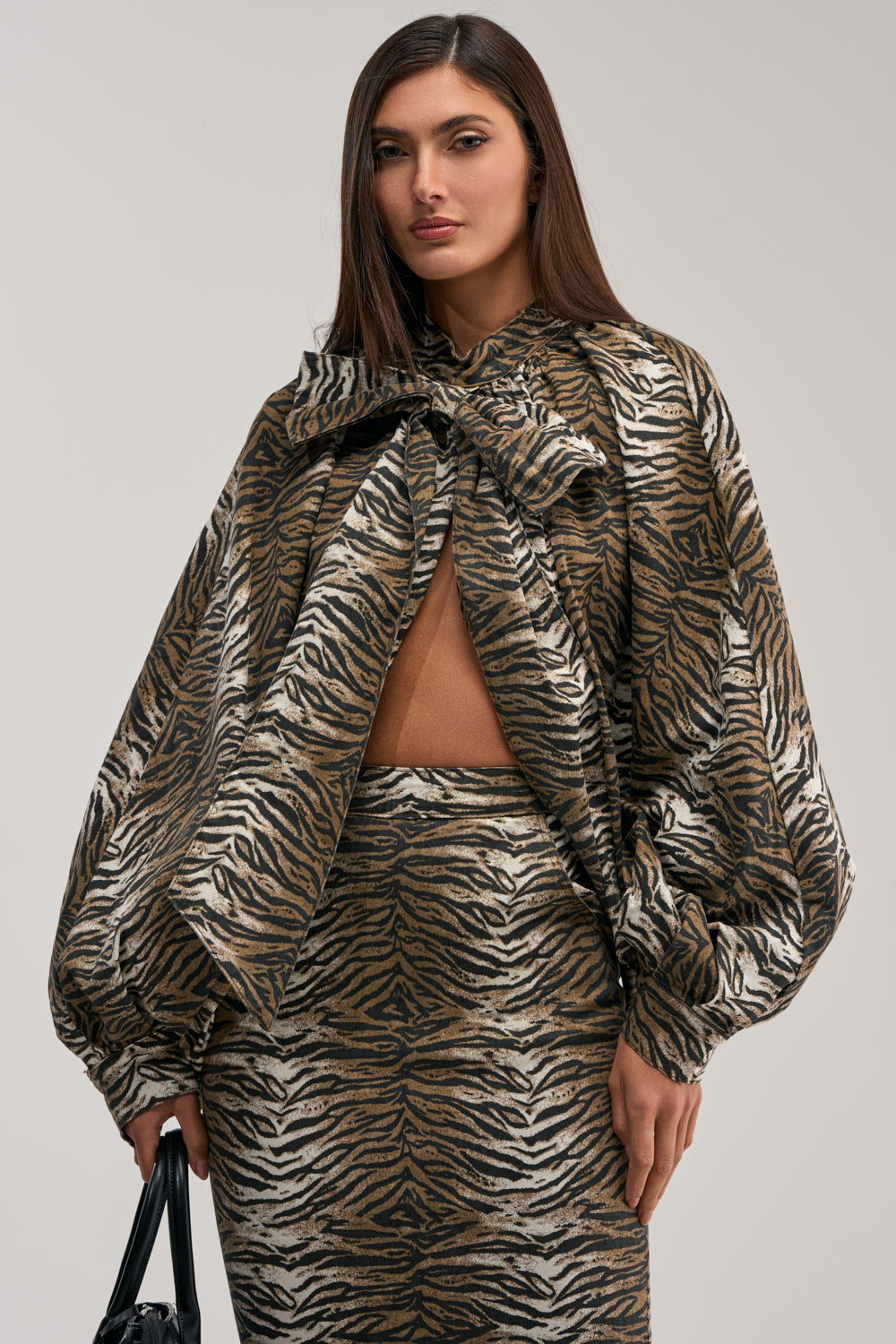 A woman with long brown hair wears the INTO THE WILD ZEBRA BLOUSE, featuring puff sleeves and an oversized neck bow, paired with a skirt. She holds a black handbag and stands against a plain light gray background.