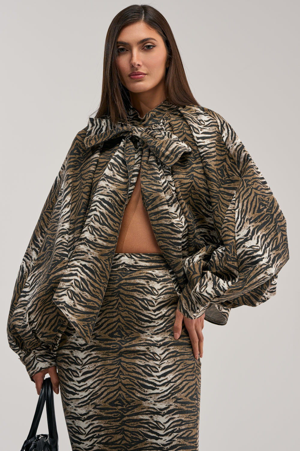 A woman models the INTO THE WILD ZEBRA BLOUSE with voluminous sleeves and an oversized bow, styled with a fitted skirt and black bag. She stands against a plain background, her long straight hair completing the statement summer look.