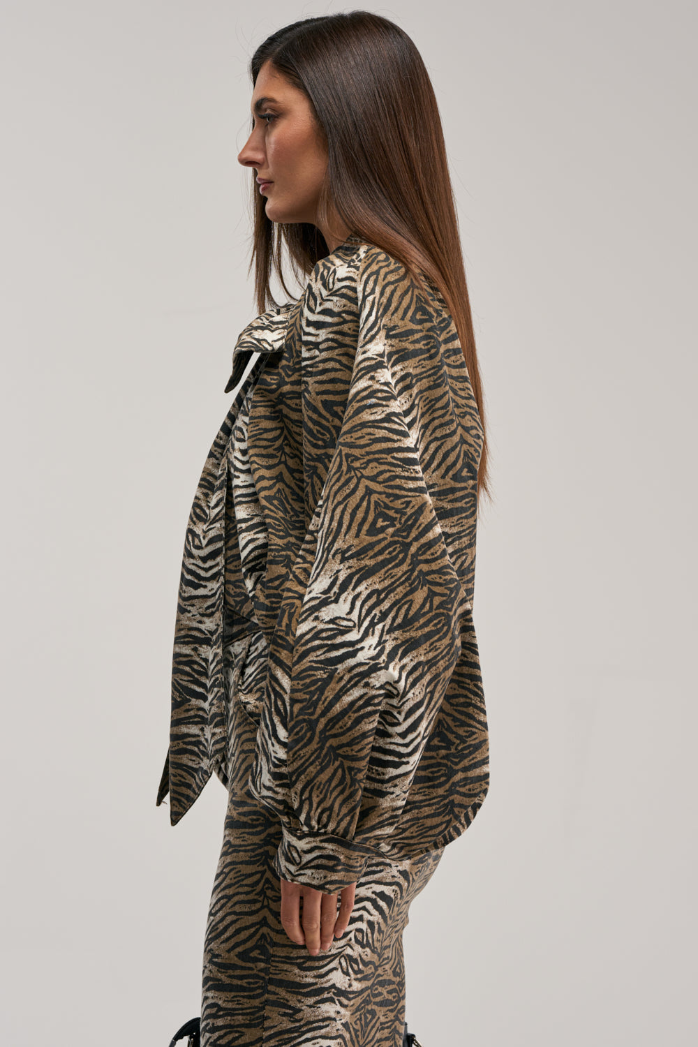 A woman with long straight brown hair stands in profile against a plain light gray background, wearing the INTO THE WILD ZEBRA BLOUSE.