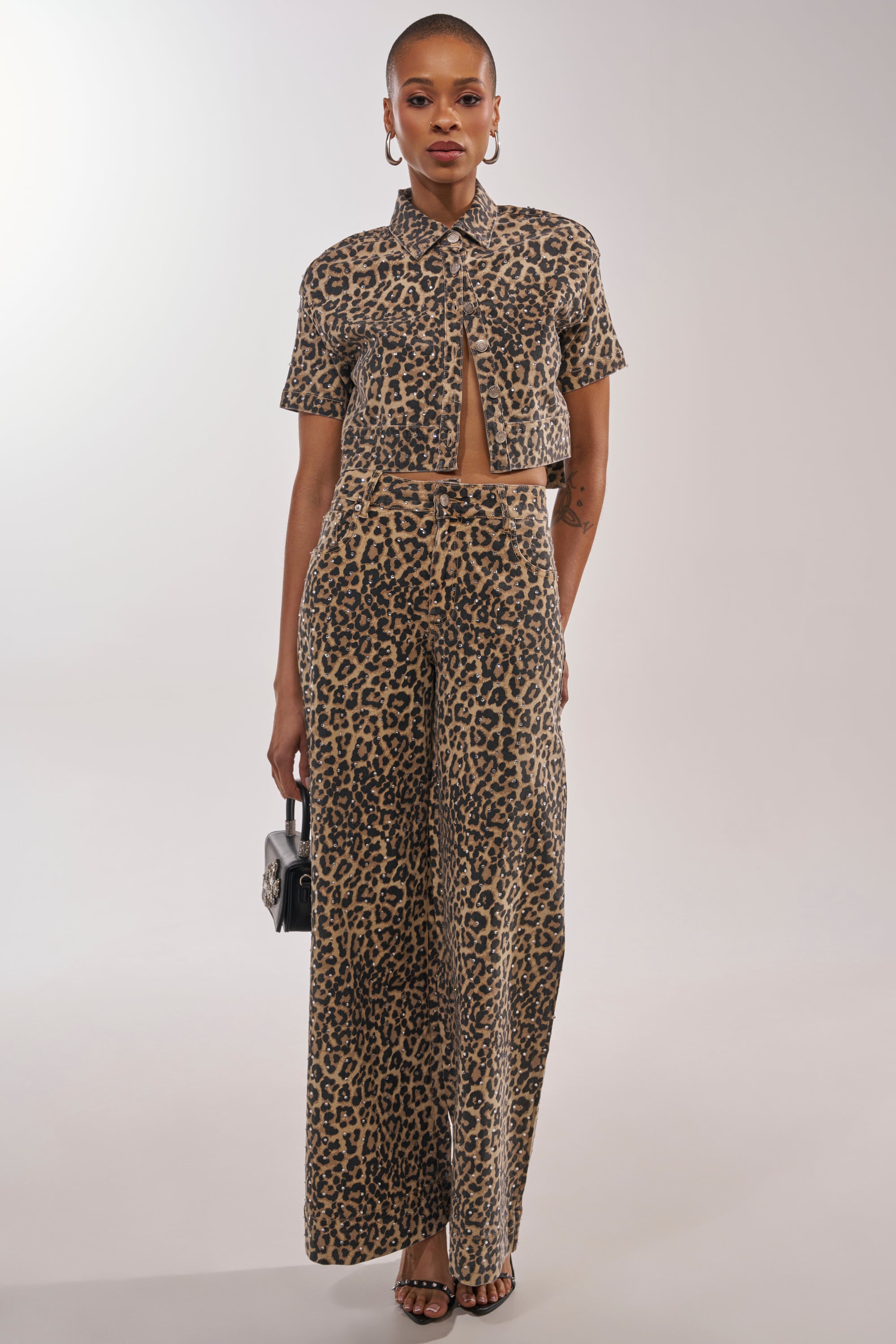 A person poses against a plain background in LEOPARD SISTA RELAXED DENIM jeans and a matching leopard print short-sleeve shirt, accessorized with hoop earrings, strappy sandals, and a small black handbag. 0