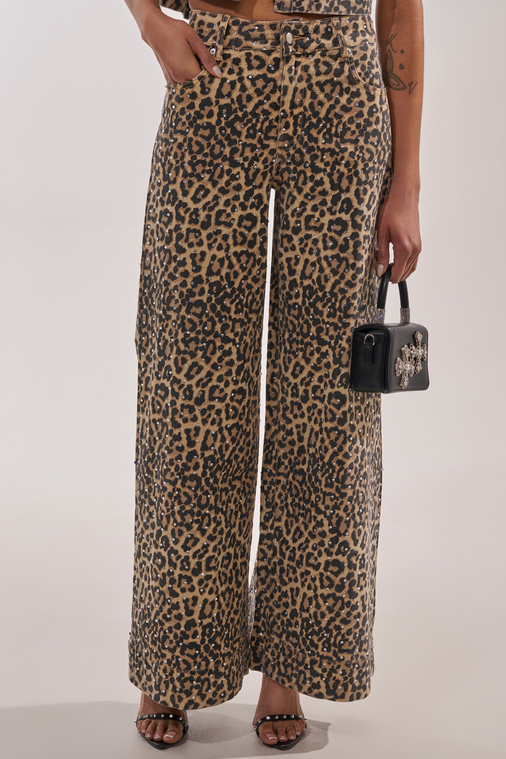 A person wears the LEOPARD SISTA RELAXED DENIM—high-waisted, wide-leg leopard print pants—paired with a small black handbag and black heeled sandals. Only the lower half is shown against a plain background. 2