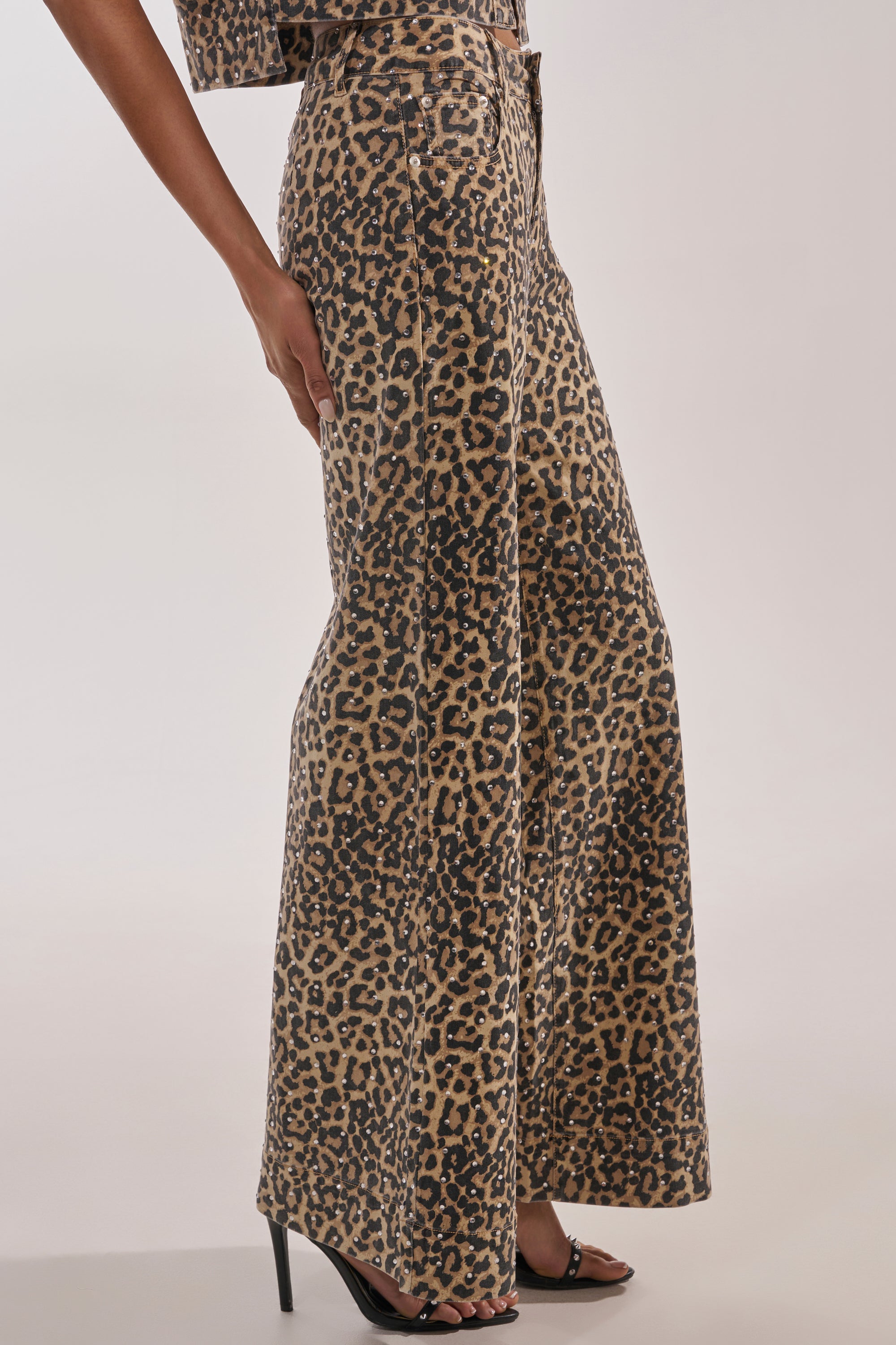 A person stands against a plain light background wearing LEOPARD SISTA RELAXED DENIM—high-waisted, wide-leg, leopard-print jeans—paired with black high-heeled sandals. 3