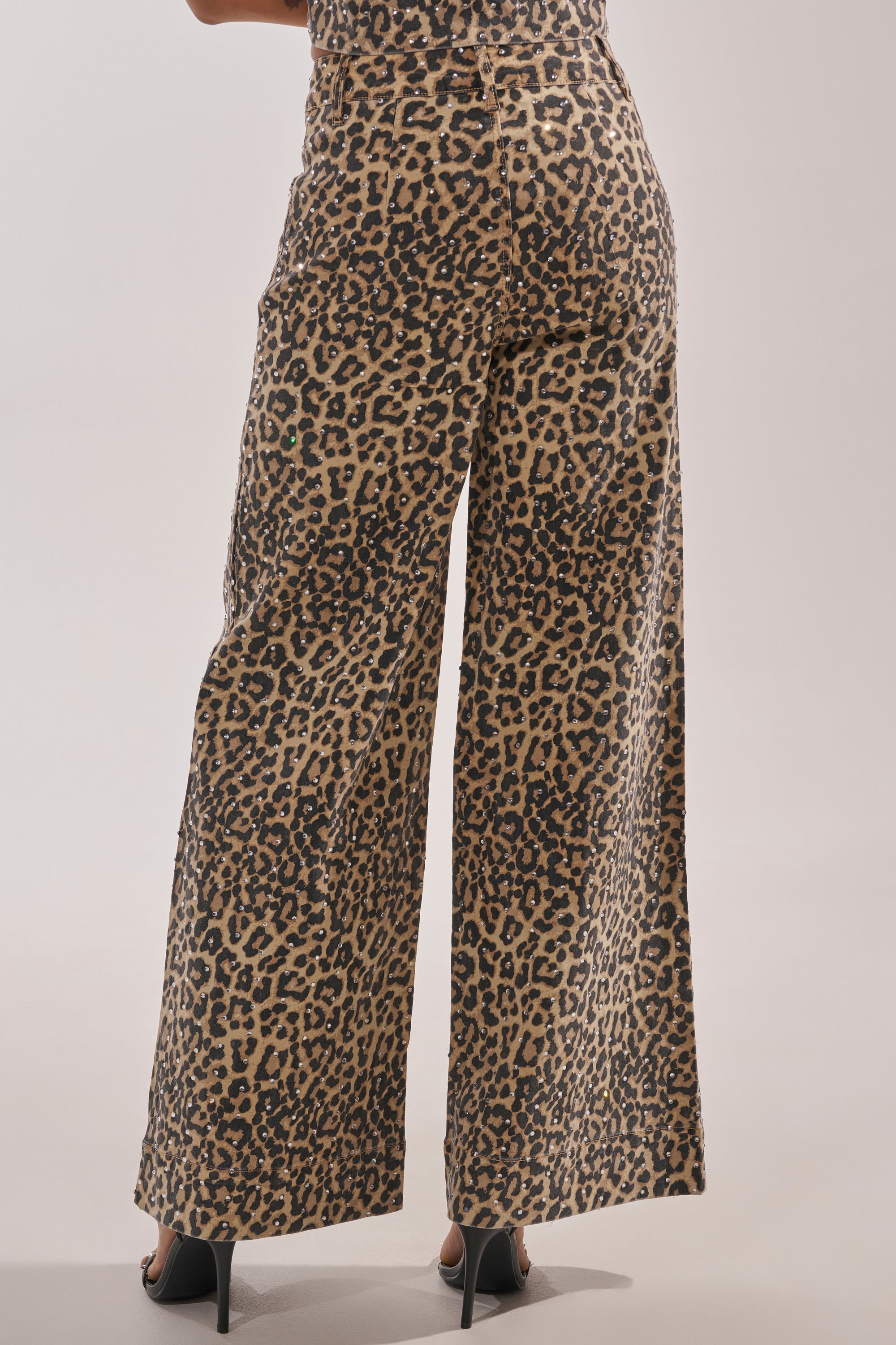 A person wears the LEOPARD SISTA RELAXED DENIM, high-waisted leopard print jeans with a wide-leg fit, teamed with black heels. The back view photo showcases the full length and bold pattern of the pants. 4