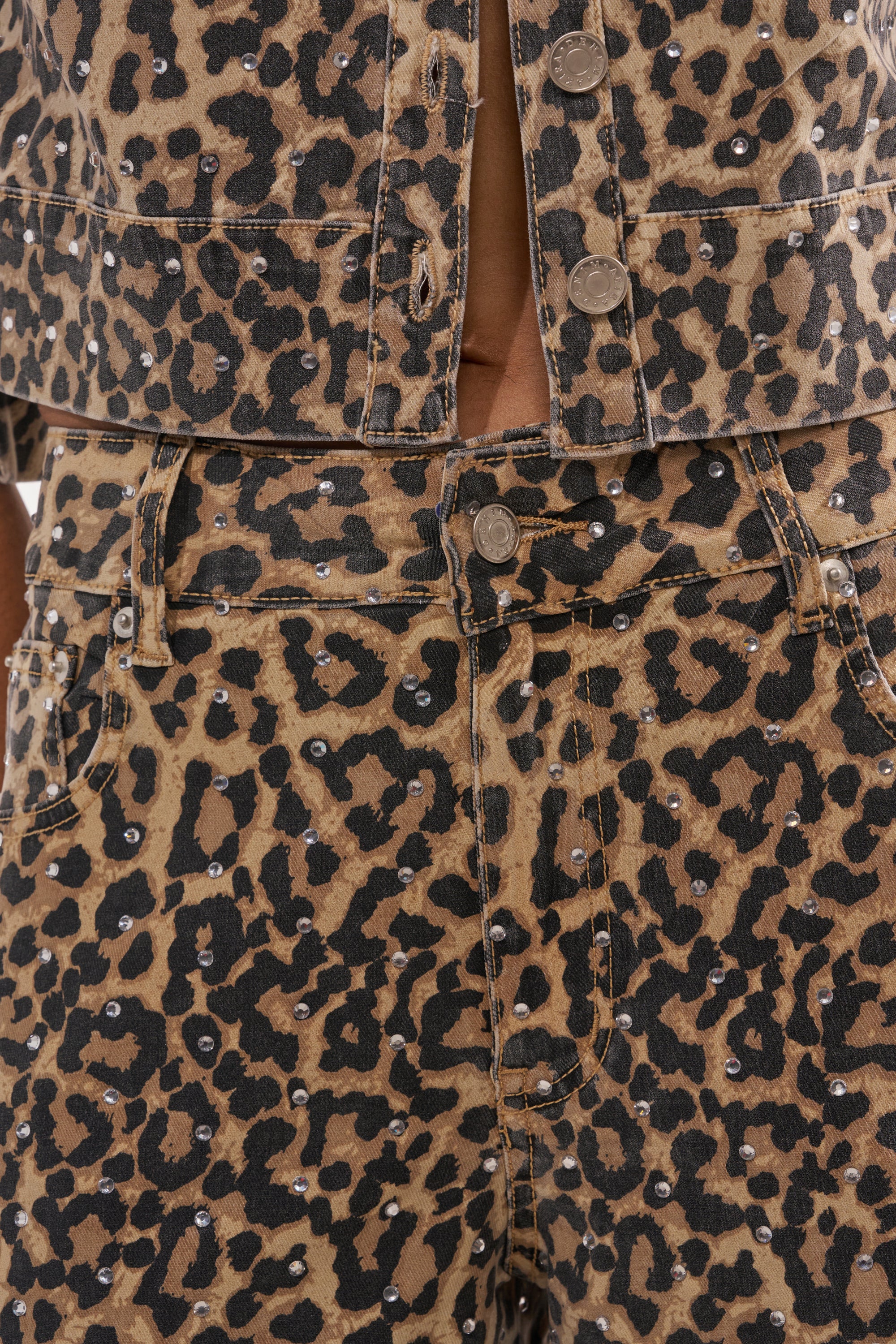 Close-up of a person wearing the LEOPARD SISTA RELAXED DENIM—leopard-print jeans and a matching jacket with silver studs. The wide-leg fit offers a modern look, shown from the torso to waist. 5