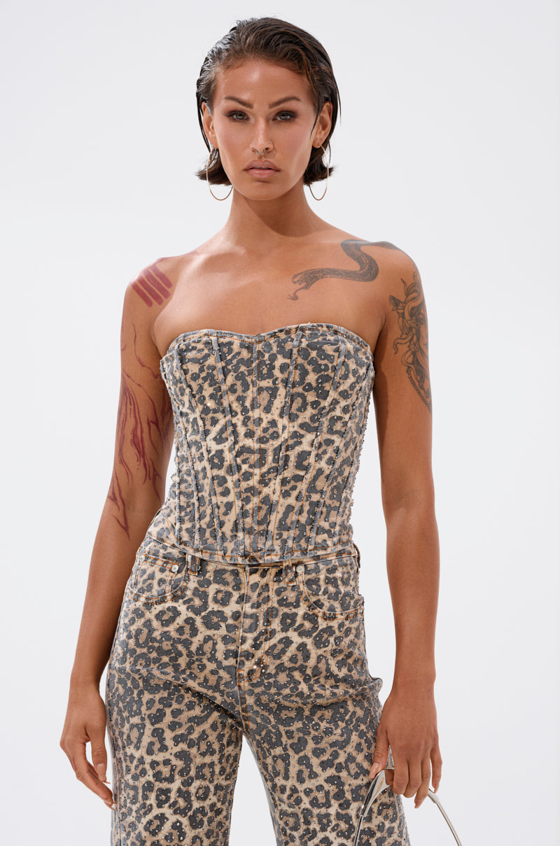 A person with short, slicked-back hair and visible arm and shoulder tattoos models the IN THE WILD CHEETAH CORSET with matching pants, standing confidently against a plain white background.