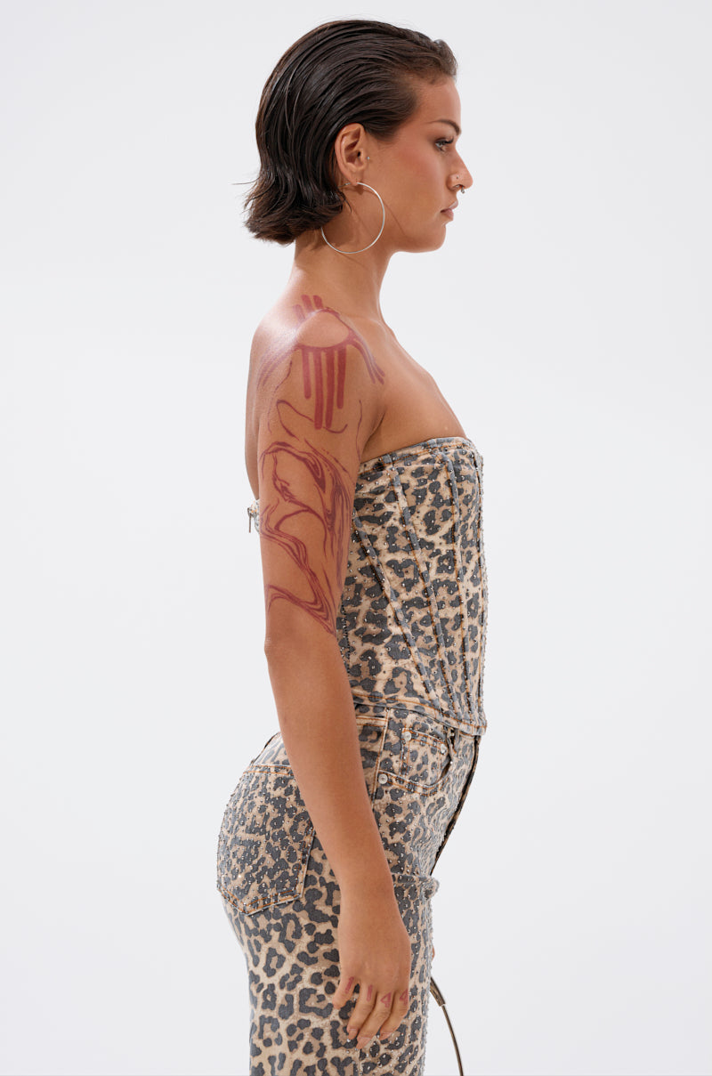 A woman with short dark hair and hoop earrings stands in profile, wearing the IN THE WILD CHEETAH CORSET. She has a bold red abstract tattoo on her upper arm, set against a plain white background.