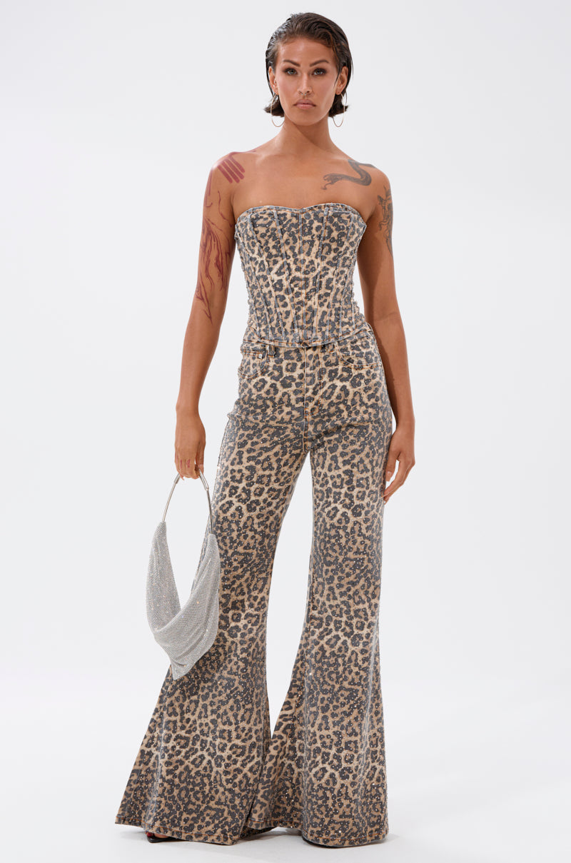 A woman with wet-look hair and visible tattoos models the IN THE WILD CHEETAH CORSET, a strapless jumpsuit with flared legs. She holds a small silver handbag and stands against a plain white background.