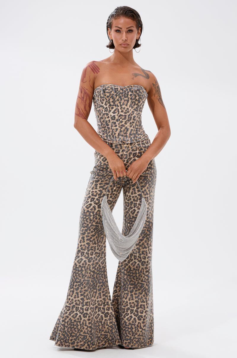 A person faces forward in the IN THE WILD CHEETAH CORSET jumpsuit with flared legs, holding a silver mesh handbag. They have wet-look hair, visible tattoos on their right arm, and stand against a plain white background.