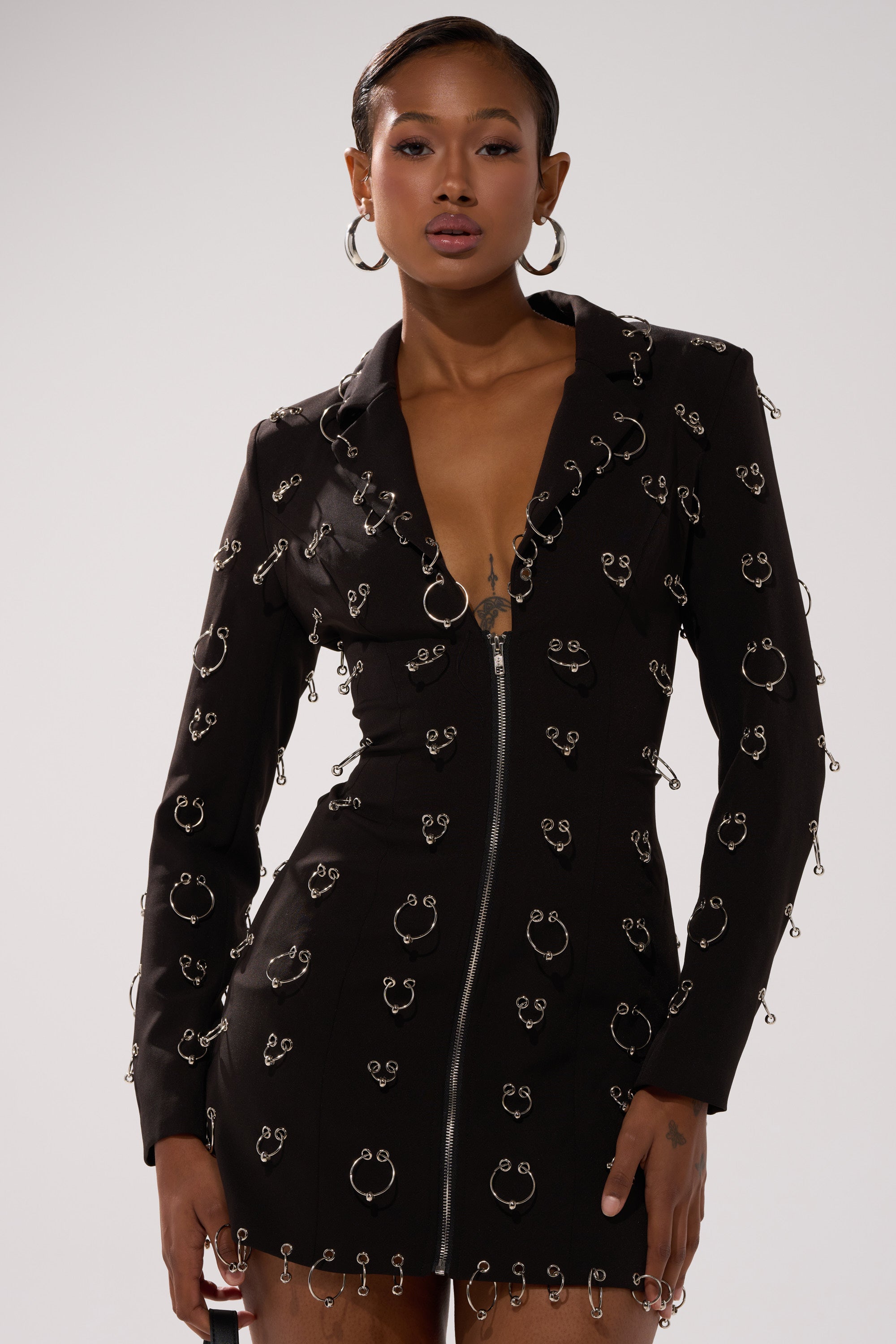 A woman models the ALL OVER PIERCING BLAZER DRESS—a tailored mini with silver hoops, safety pins, a deep V-neckline, and long sleeves. She pairs it with large hoop earrings and slicked-back hair for an edgy look.