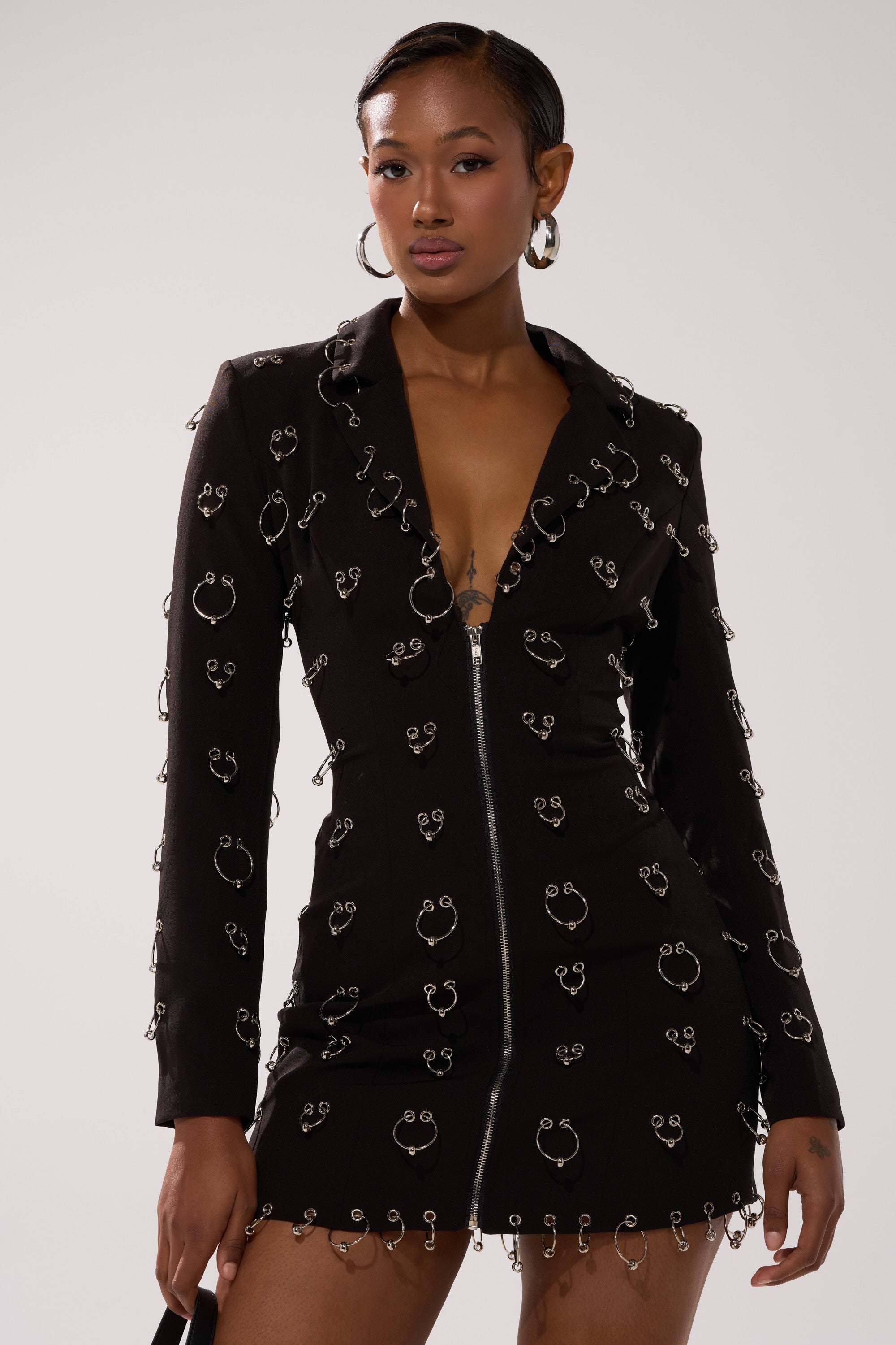 A woman models the ALL OVER PIERCING BLAZER DRESS, a tailored mini dress with silver rings and a front zipper. She has short, sleek hair, wears hoop earrings, and holds a small black accessory against a plain light background.