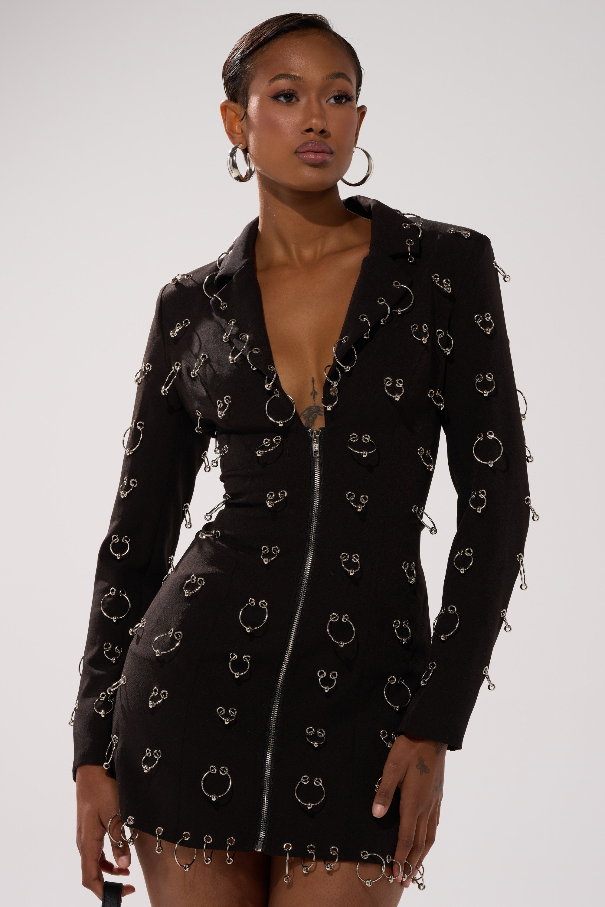 A person wearing the ALL OVER PIERCING BLAZER DRESS, a black long-sleeved mini dress with silver safety pin details, styled with large hoop earrings and posing against a plain light background.