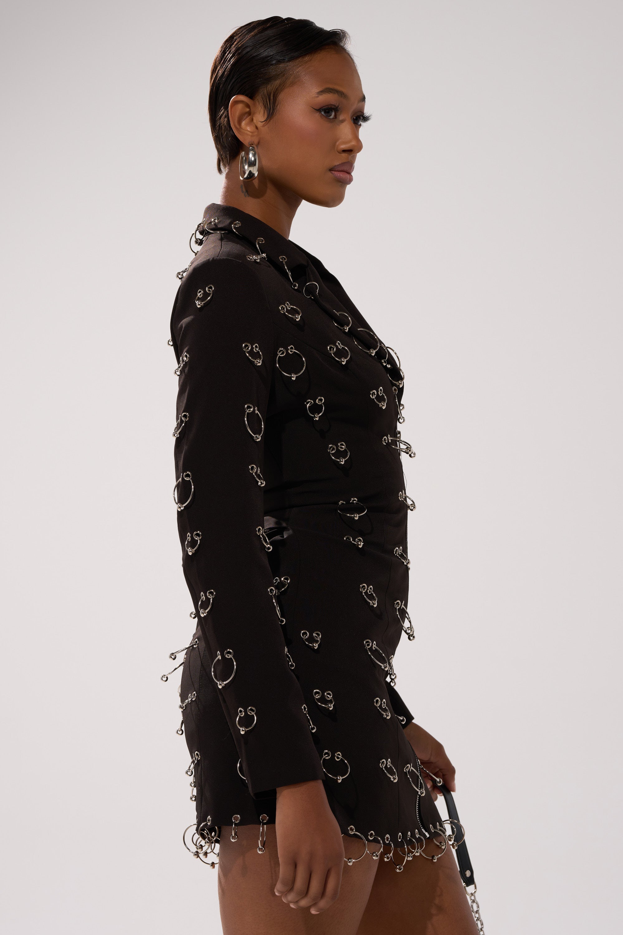 A woman in profile models the ALL OVER PIERCING BLAZER DRESS—black, featuring metallic rings and embellishments. She wears hoop earrings and has short, slicked-back hair, standing before a plain light background.
