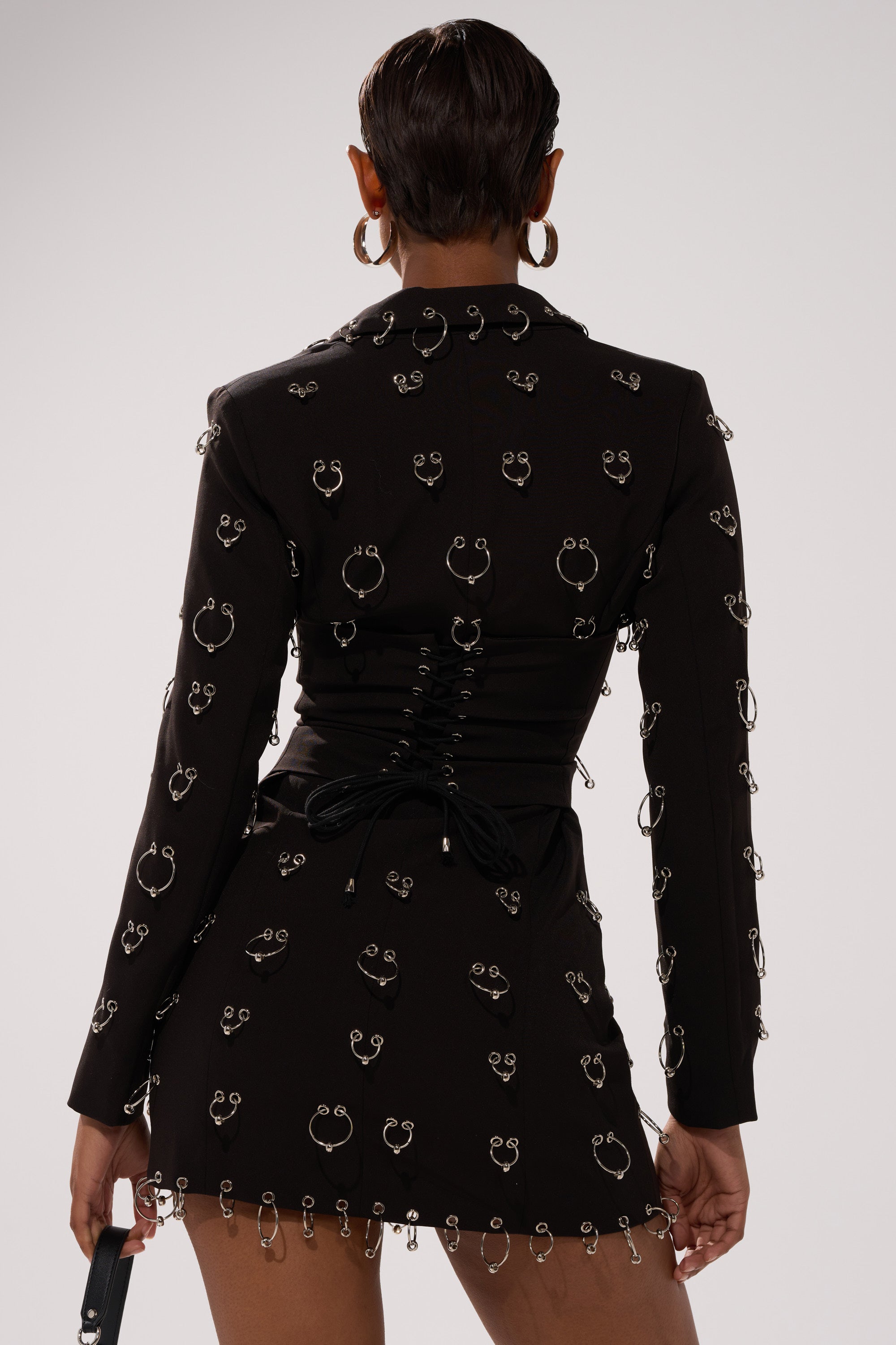 A person with short hair models the ALL OVER PIERCING BLAZER DRESS, featuring silver ring accents and a corset lace-up back, against a plain light background.