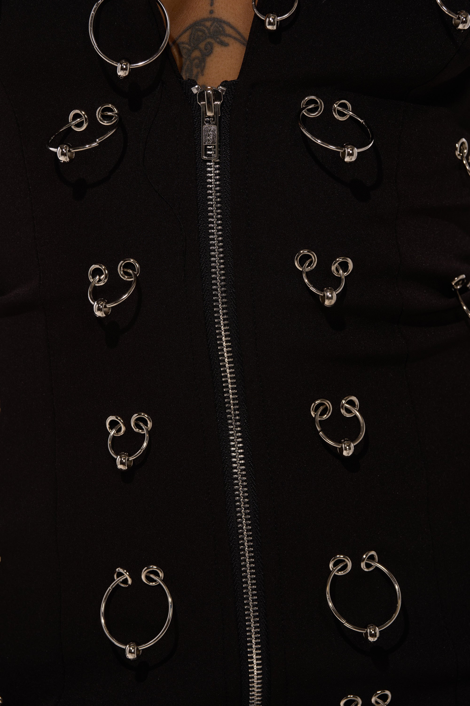 Close-up of the ALL OVER PIERCING BLAZER DRESS, a black tailored mini dress with a central zipper and multiple silver circular rings. A hint of tattoo peeks out at the top, adding edge to this standout piece.