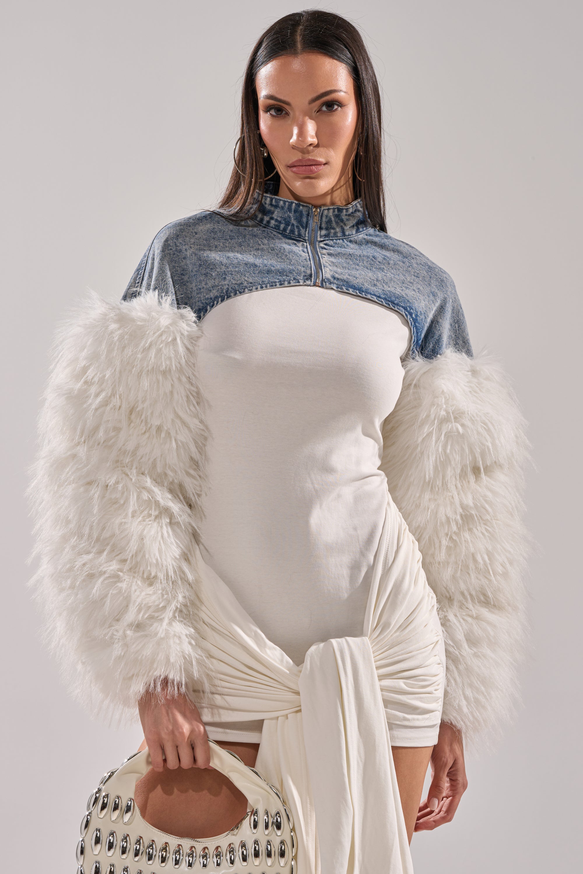 A woman poses in the CELESTE FUR SLEEVE BOLERO—a white dress with fluffy, feathered sleeves and a denim bolero upper bodice. She holds a white handbag with metallic accents, her long hair straight, against a plain light background. 0
