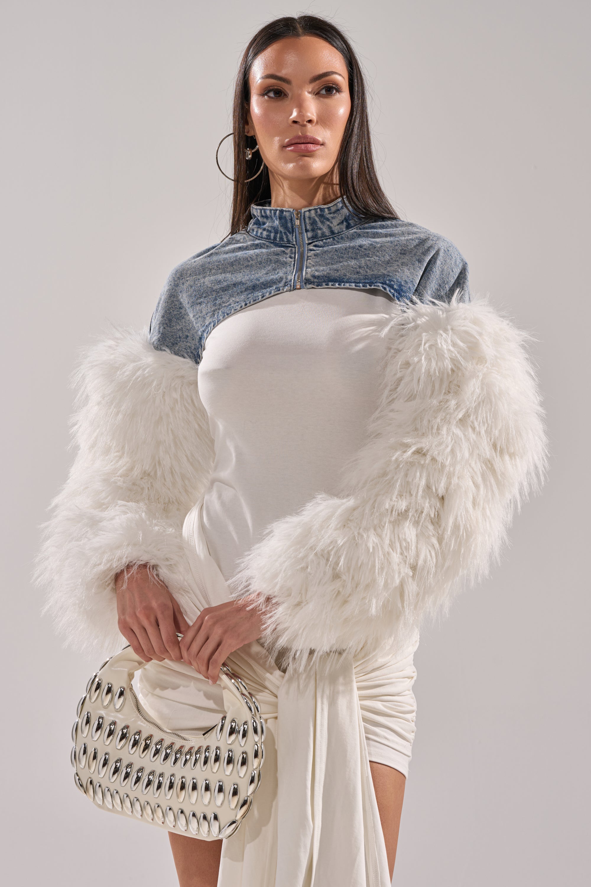 A woman poses in the CELESTE FUR SLEEVE BOLERO, featuring dramatic long white faux fur sleeves and a cropped denim cut. She accessorizes with a silver oval-studded handbag and large hoop earrings, standing against a plain light background. 1