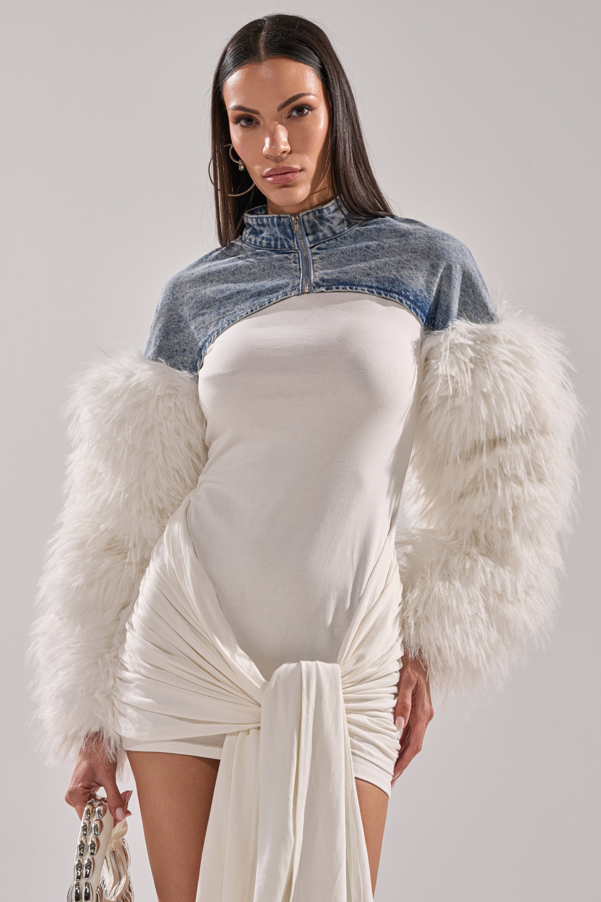 A woman styles the CELESTE FUR SLEEVE BOLERO—a cropped denim jacket with oversized white faux fur sleeves—over a fitted white ruched dress, accessorizing with a chain-handle bag for a chic, confident look. 2