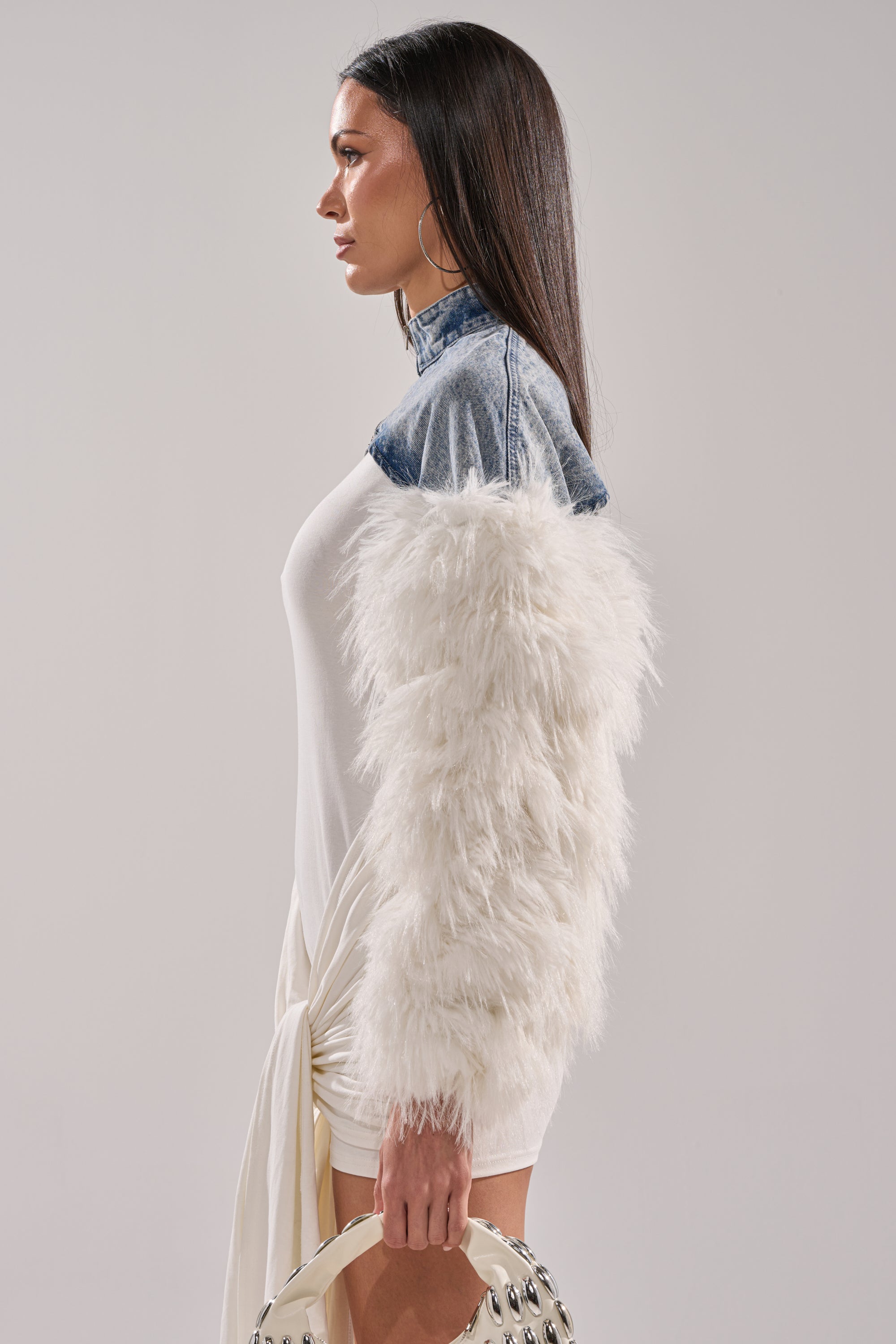 A woman stands in profile wearing the CELESTE FUR SLEEVE BOLERO with a denim collar over a white dress, holding a cream-colored bag with metal details. She has long, straight dark hair and large hoop earrings. 4