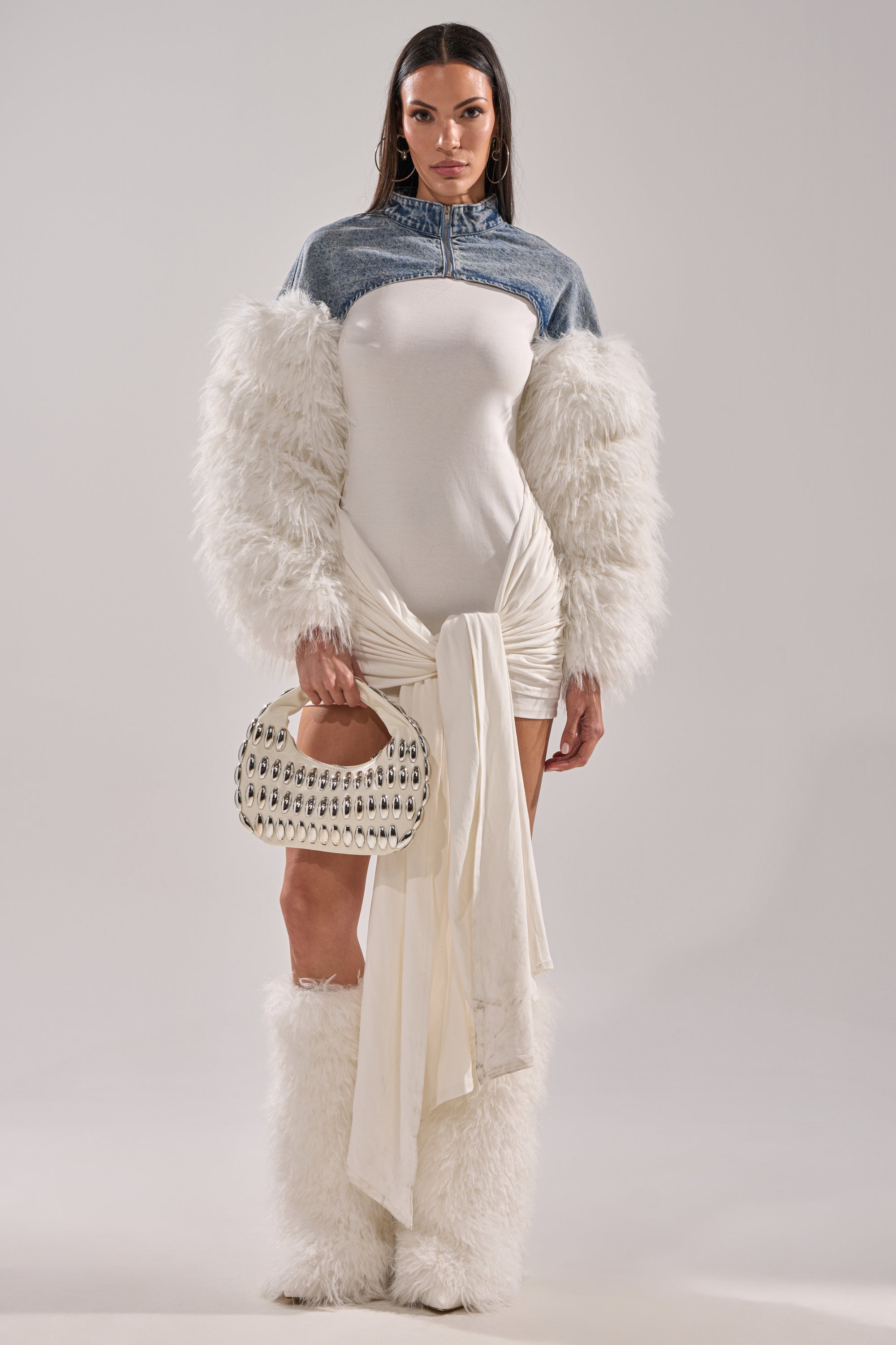 A woman faces forward in a white dress with a ruched skirt, straight dark hair, and holds a studded handbag. She layers her look with the CELESTE FUR SLEEVE BOLERO and matching furry boots against a plain background. 6
