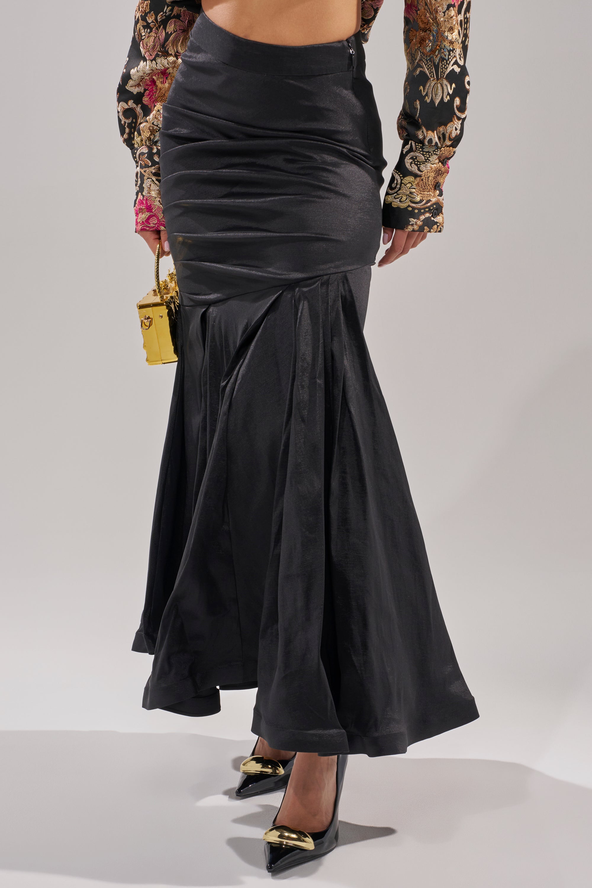A person wearing the QUEEN OF IT ALL MAXI SKIRT in black with a ruched, mermaid hemline, paired with black heels featuring gold tips and a small gold purse. The top is colorfully floral-embroidered—ideal for formal events.