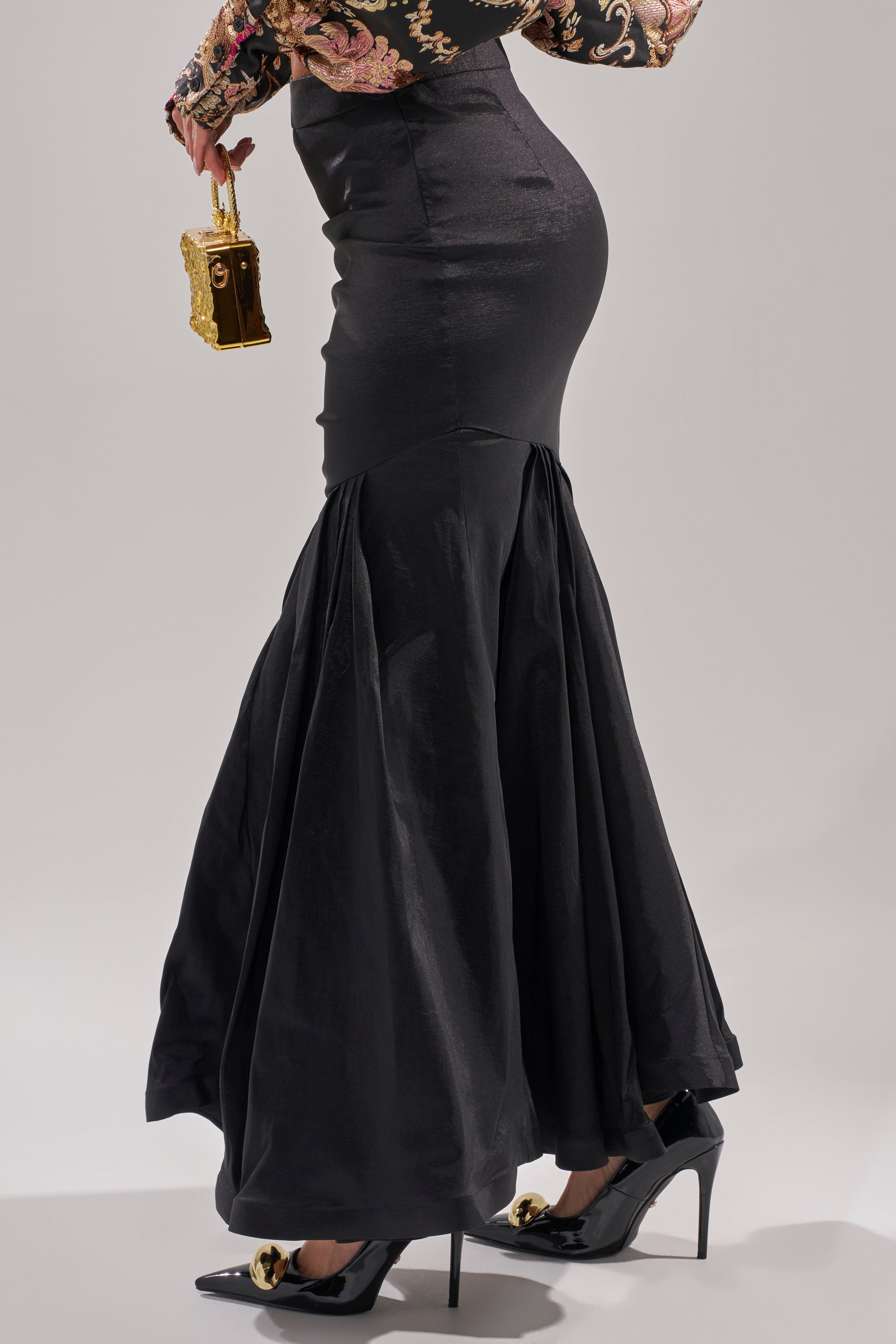 Wearing the QUEEN OF IT ALL MAXI SKIRT with a mermaid hemline, black pointed-toe heels with gold accents, and carrying an ornate handbag—shown waist down against a plain background—an elegant look for formal occasions.