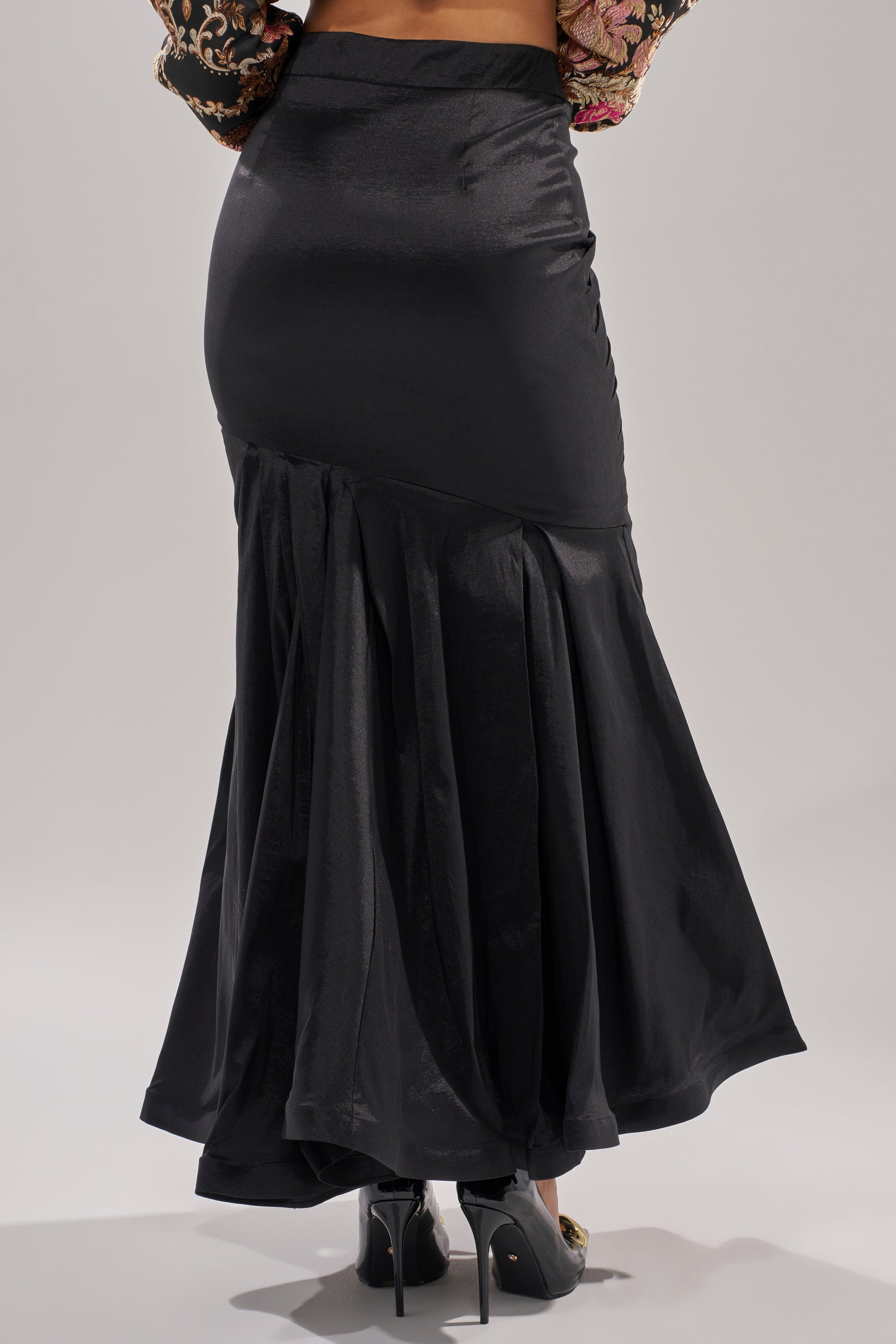 The QUEEN OF IT ALL MAXI SKIRT, a long black satin maxi with a mermaid hem, is styled with black high heels and shown from the back against a plain light background—ideal for formal occasions.