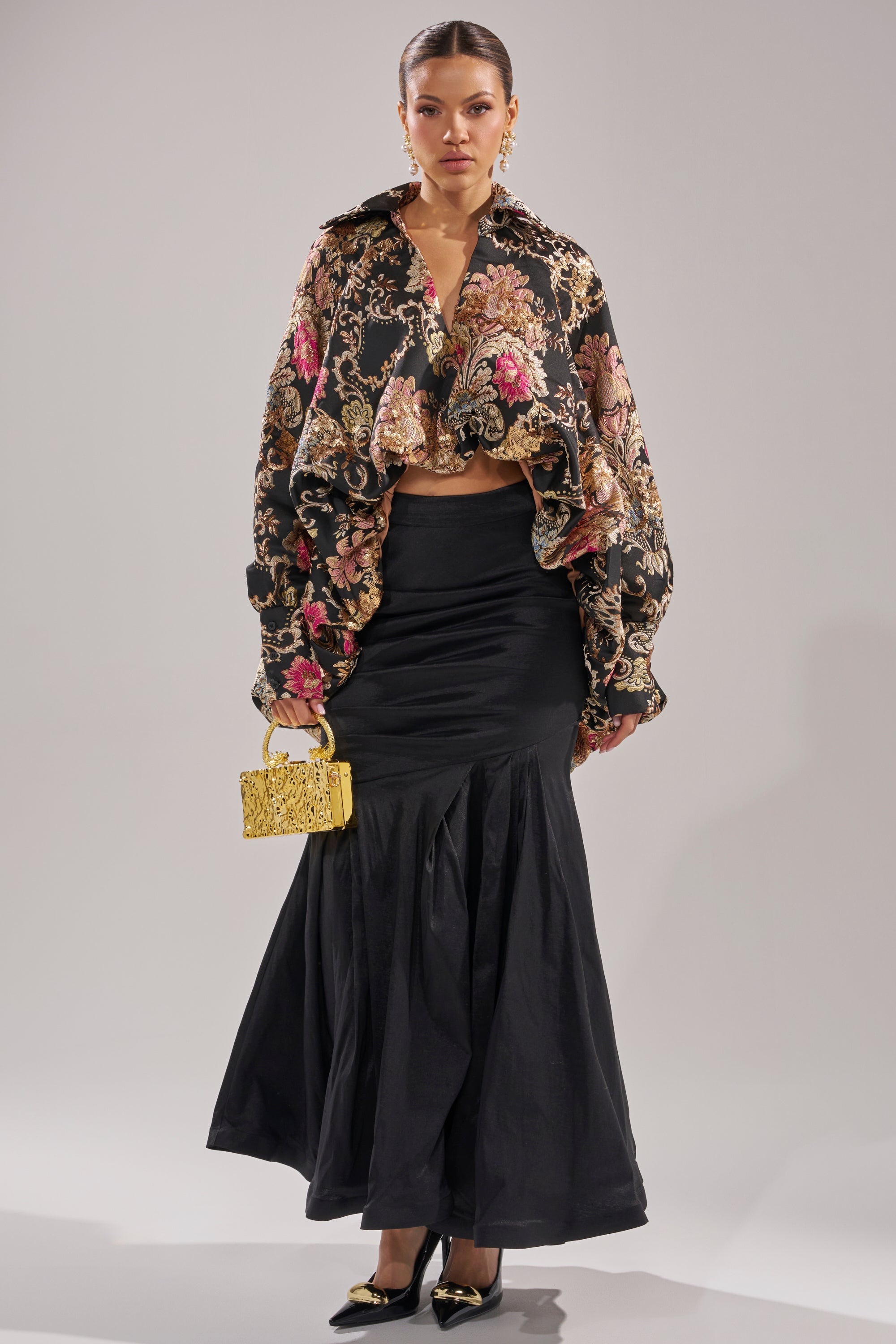 A woman faces forward in a floral ruffled blouse, voluminous sleeves, gold earrings, black pointed heels, an ornate yellow clutch, and the QUEEN OF IT ALL MAXI SKIRT with a mermaid hem—an elegant look for formal events.