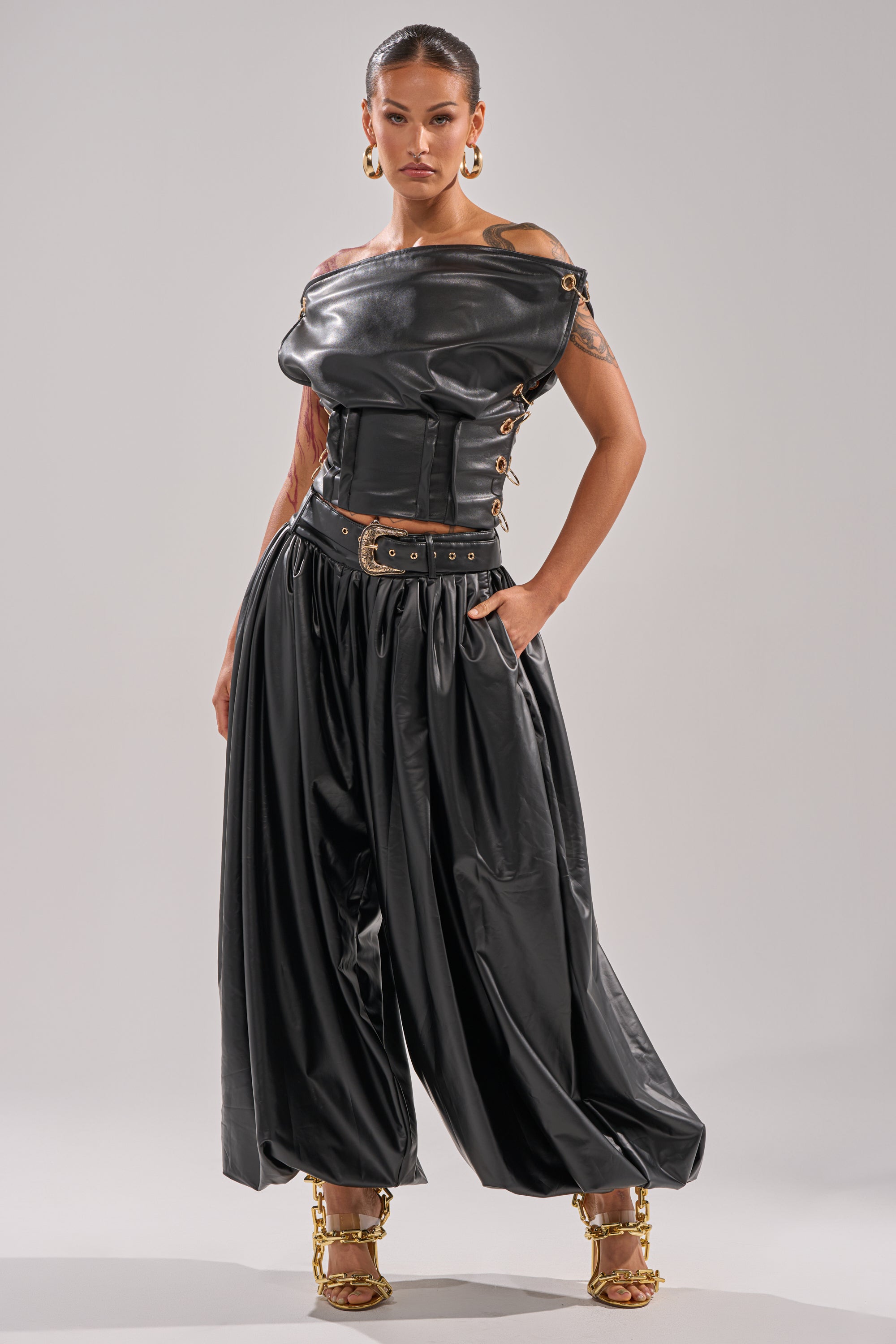 A woman poses confidently in wide-legged ZONA BALLOON LEATHER TROUSERS and a black off-shoulder leather corset top, accessorized with chunky gold jewelry and chain-detailed heels, set against a plain light gray background.