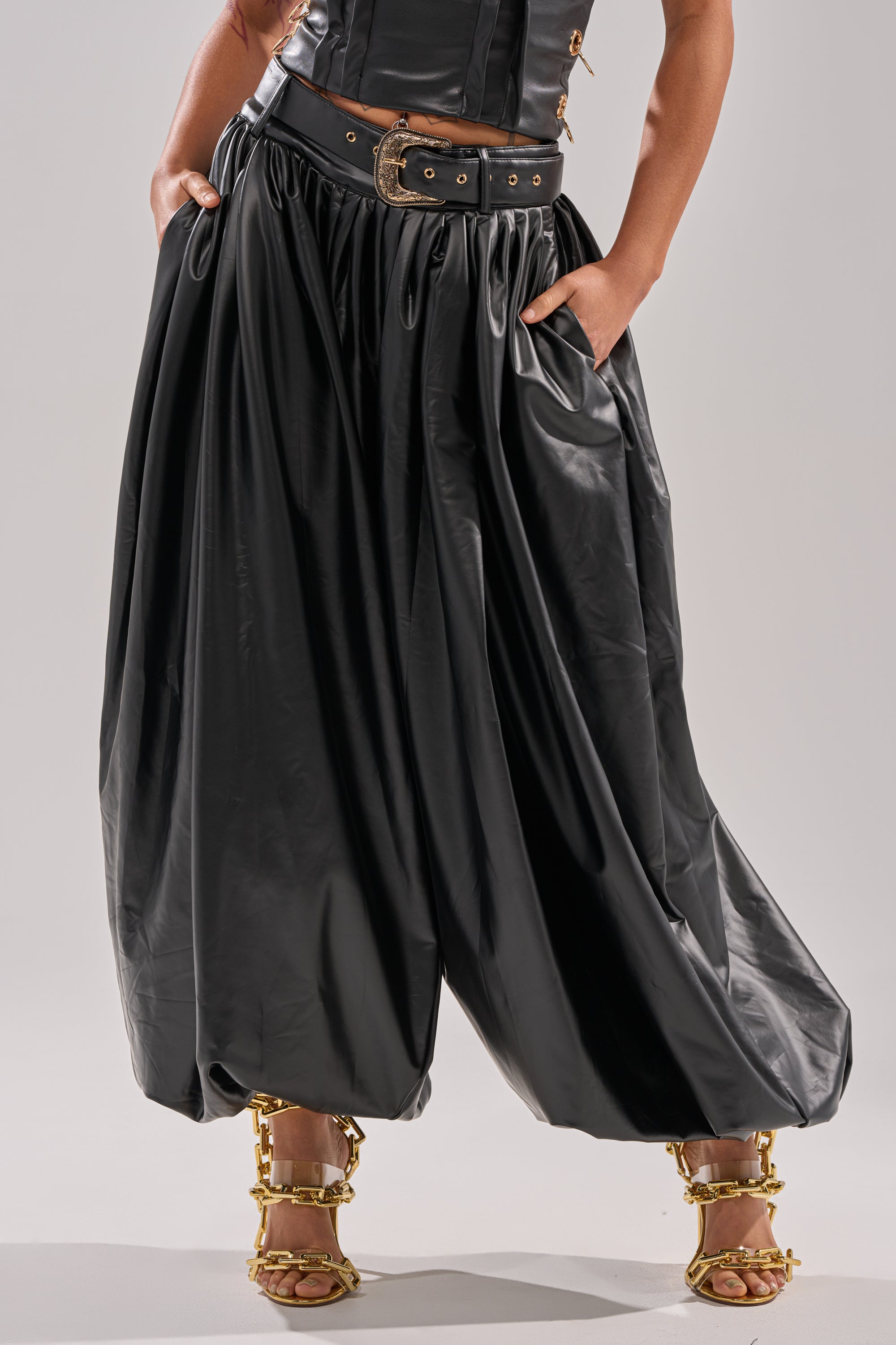 Wearing ZONA BALLOON LEATHER TROUSER in shiny black with a black belt and gold buckle, paired with gold chain-strap heels. Hands are tucked in the trouser pockets; only part of the upper body is visible.