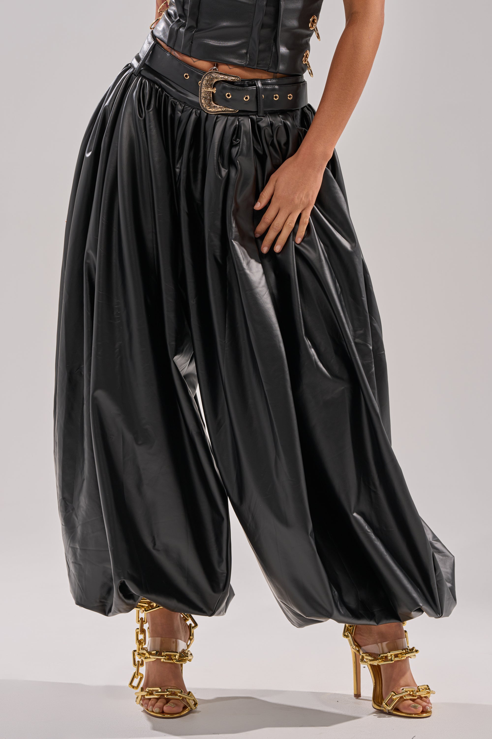 Wearing ZONA BALLOON LEATHER TROUSER, a black corset top, a wide black belt with gold buckle, and gold high-heeled sandals with chain accents, a person stands against a plain light background.